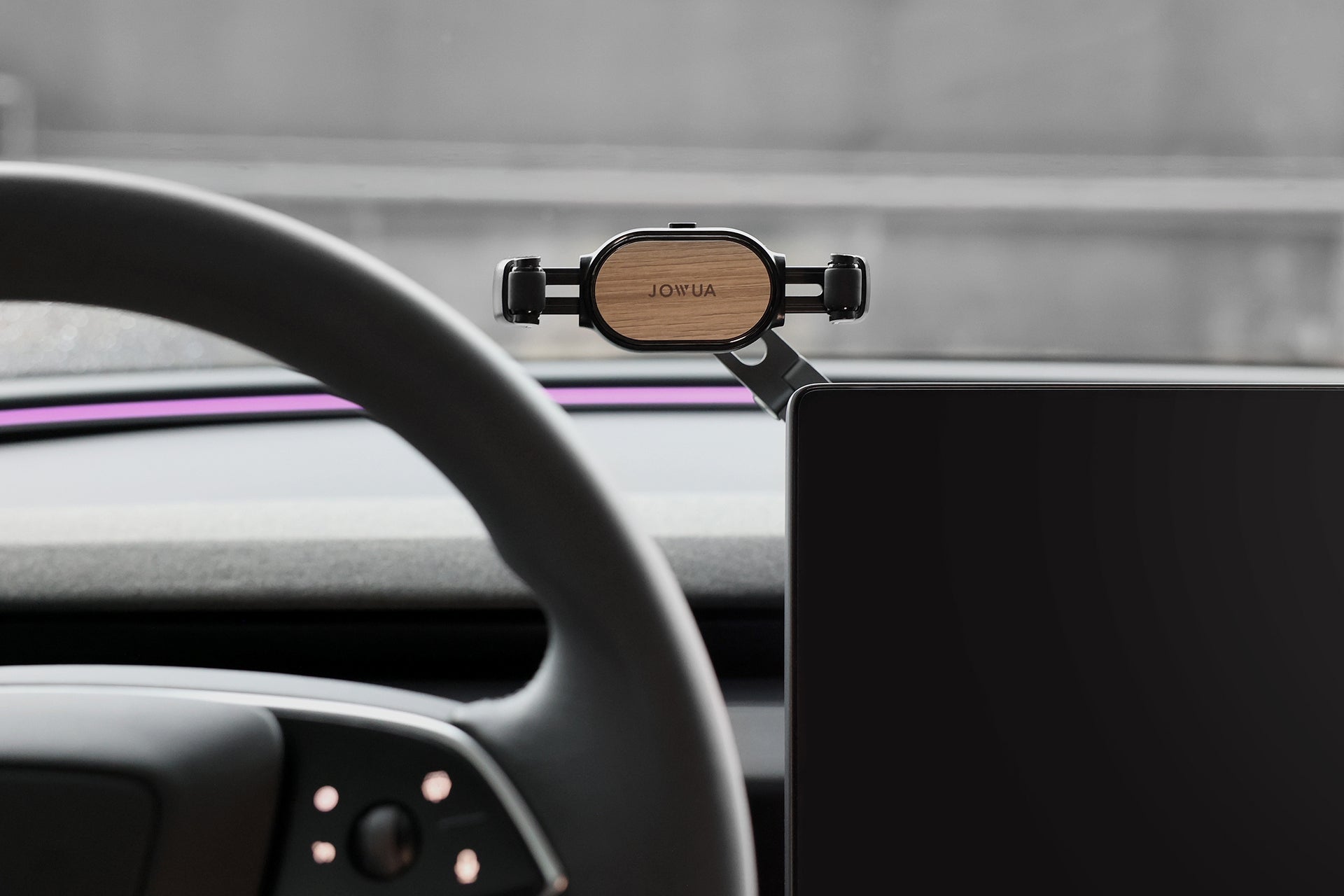 A car interior features JOWUA's 6D Invisible Foldaway Car Mount (Universal) on the dashboard. It combines durability with a wooden back and adjustable angles for a secure grip. The sleek design complements the visible steering wheel and dashboard screen, enhancing functionality and style.