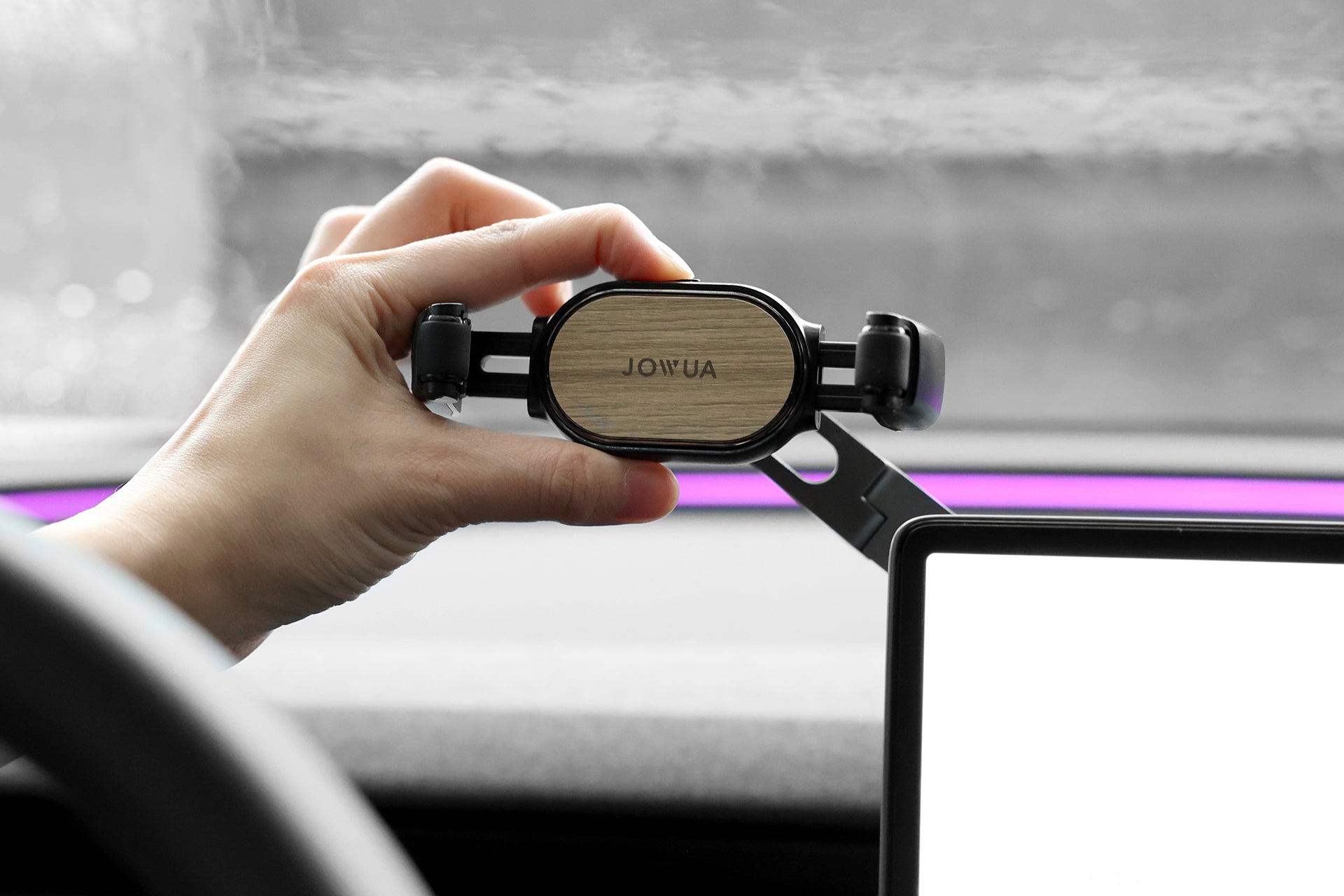 A person is installing the JOWUA 6D Invisible Foldaway Car Mount (Universal) with a sleek wooden accent on their dashboard. Made from durable material, it allows adjustable angles for optimal viewing, enhancing the modern car interior with blurred windshields and side screens.