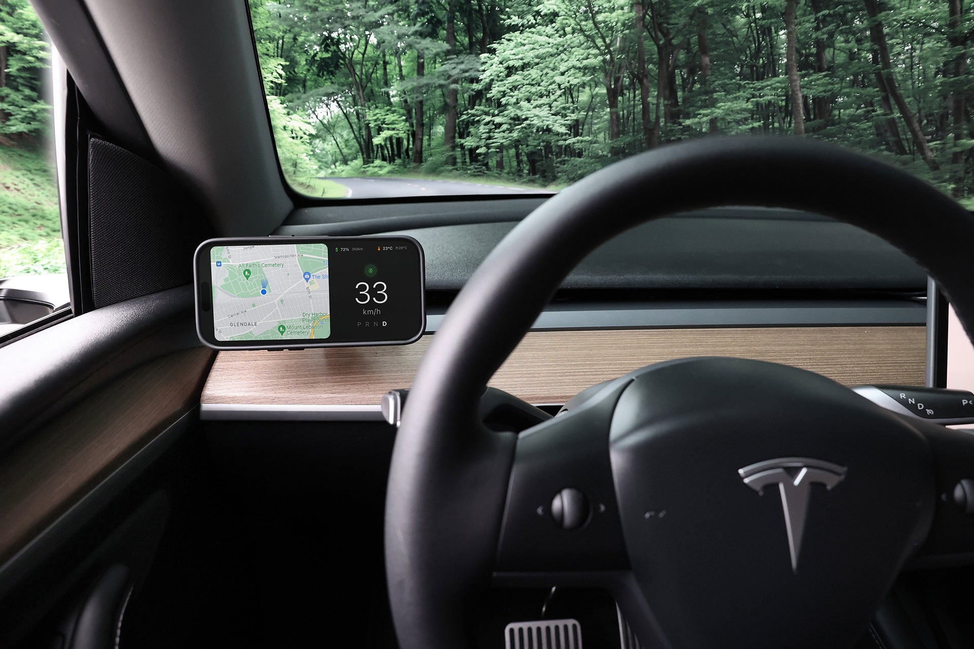 View from the driver’s seat of a Tesla shows the steering wheel, dashboard, and digital display. A JOWUA Active Cooling MagSafe Car Mount for Air Vents (Model 3/Y 2018-2024) is securely attached while driving down a tree-lined road.