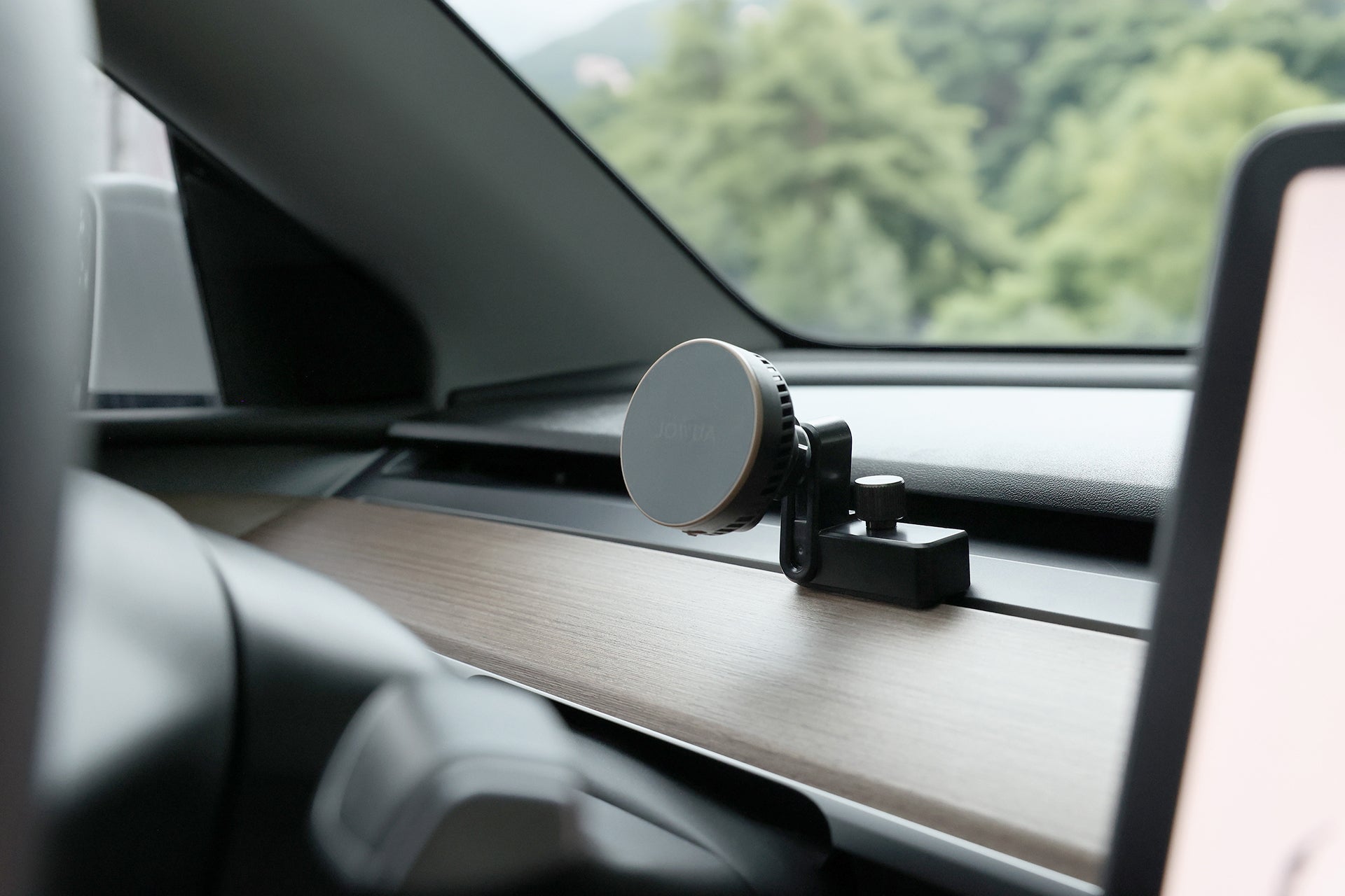 A close-up of the JOWUA Active Cooling MagSafe Car Mount for Air Vents (Model 3/Y 2018-2024) attached to a car’s wooden dashboard, with greenery visible through the windshield in the background.