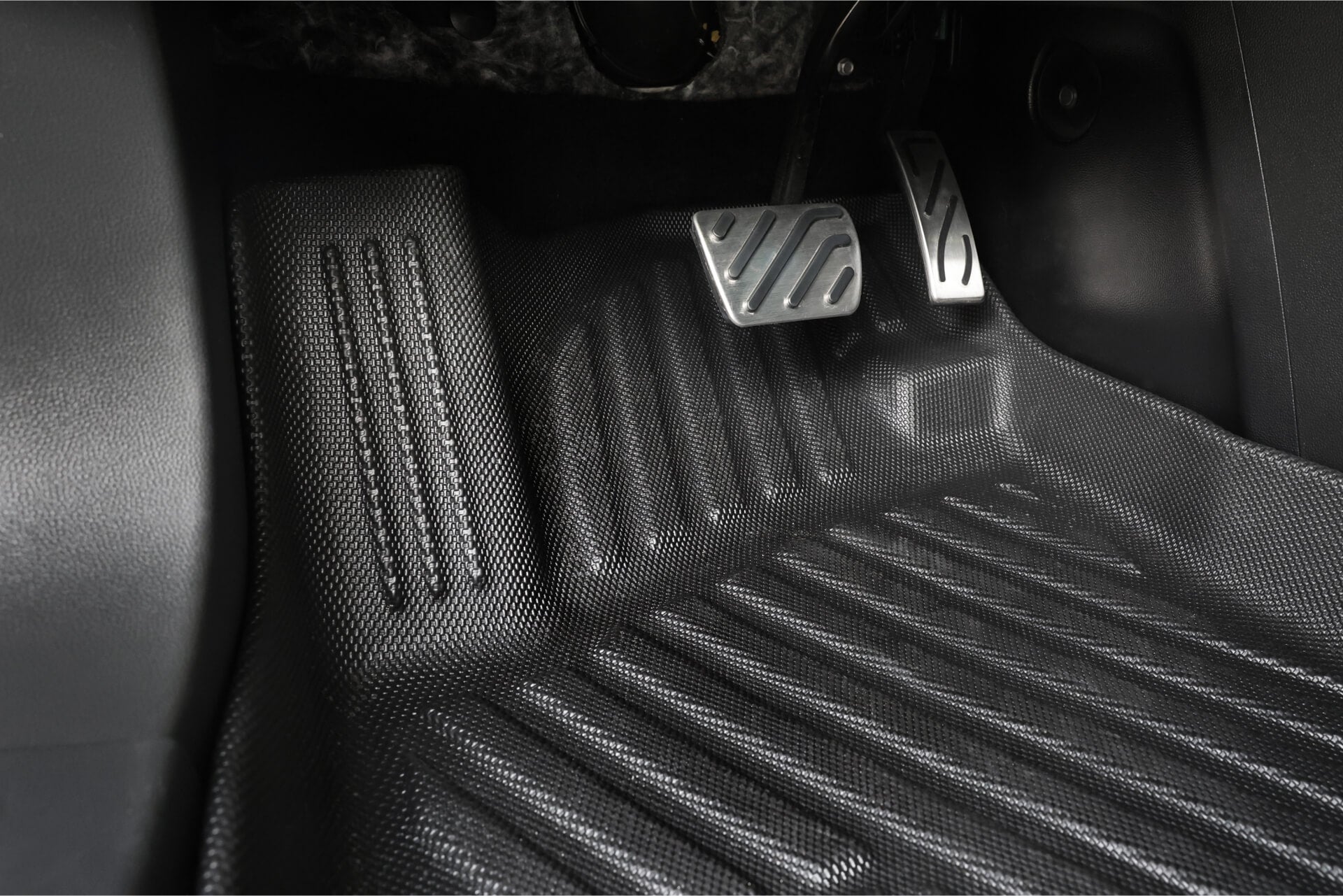 Close-up view of a car footwell featuring the JOWUA All Weather Floor Liner for MG MG4 and two metallic pedals (brake and accelerator) with patterned grip surfaces for enhanced traction.