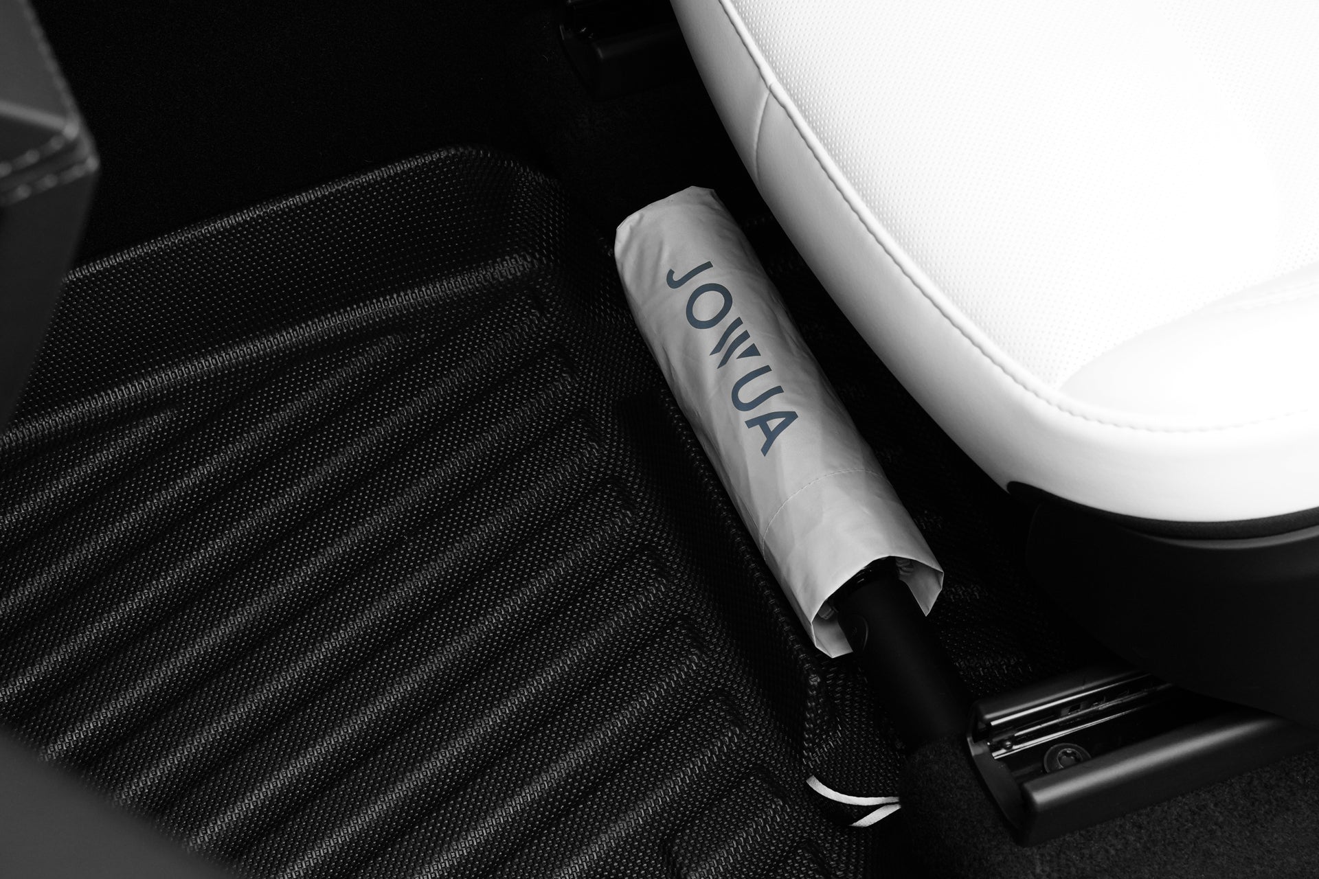 A white umbrella with "JOWUA" printed on it lies on the floor between a Tesla Model Y's white seat and the Model Y (Juniper 2025+) All-Weather Floor Liners Combo by JOWUA.