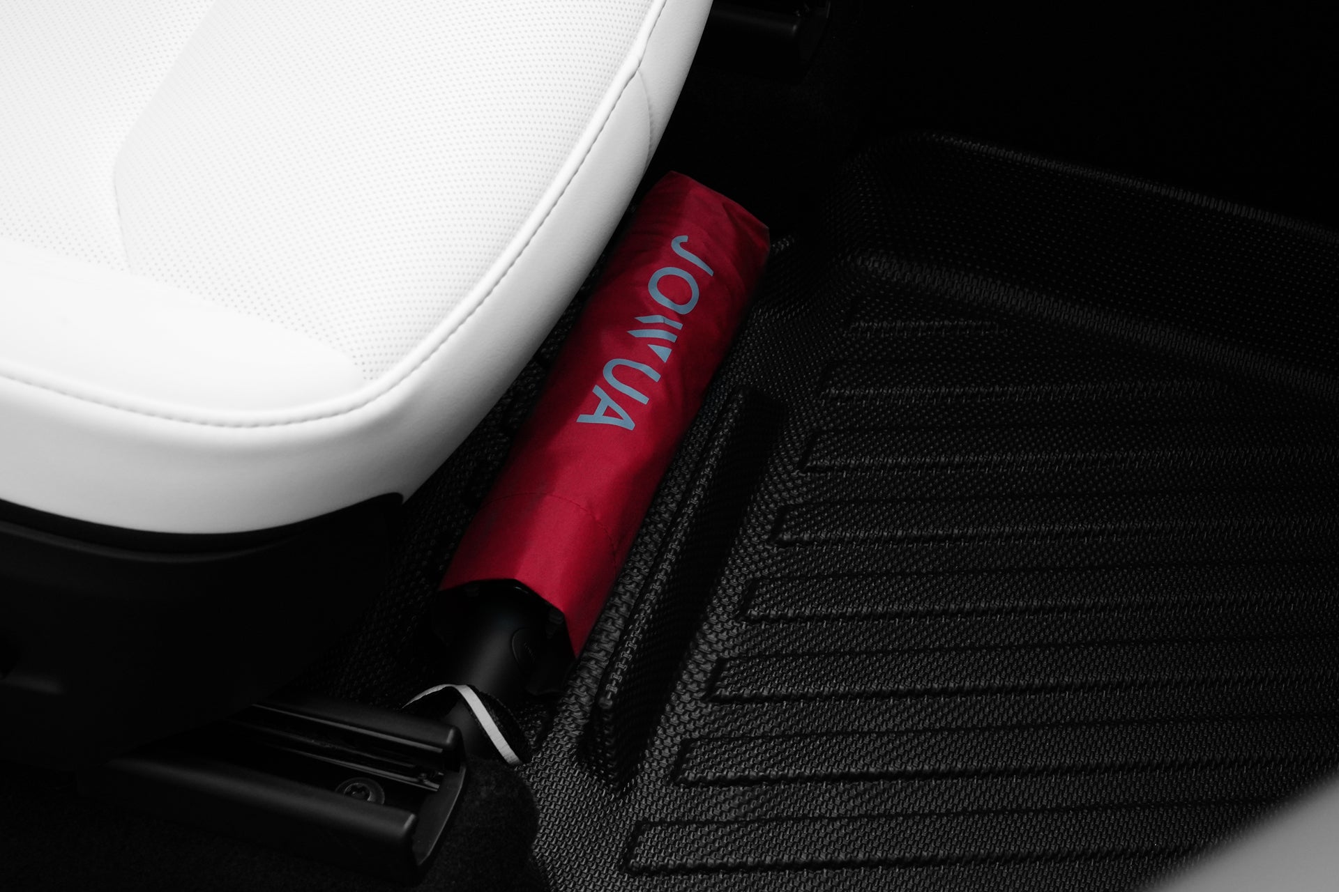 A red JOWUA umbrella sits near the edge of a white car seat, resting on Model Y (2025+) All-Weather Floor Liners Combo inside the vehicle.