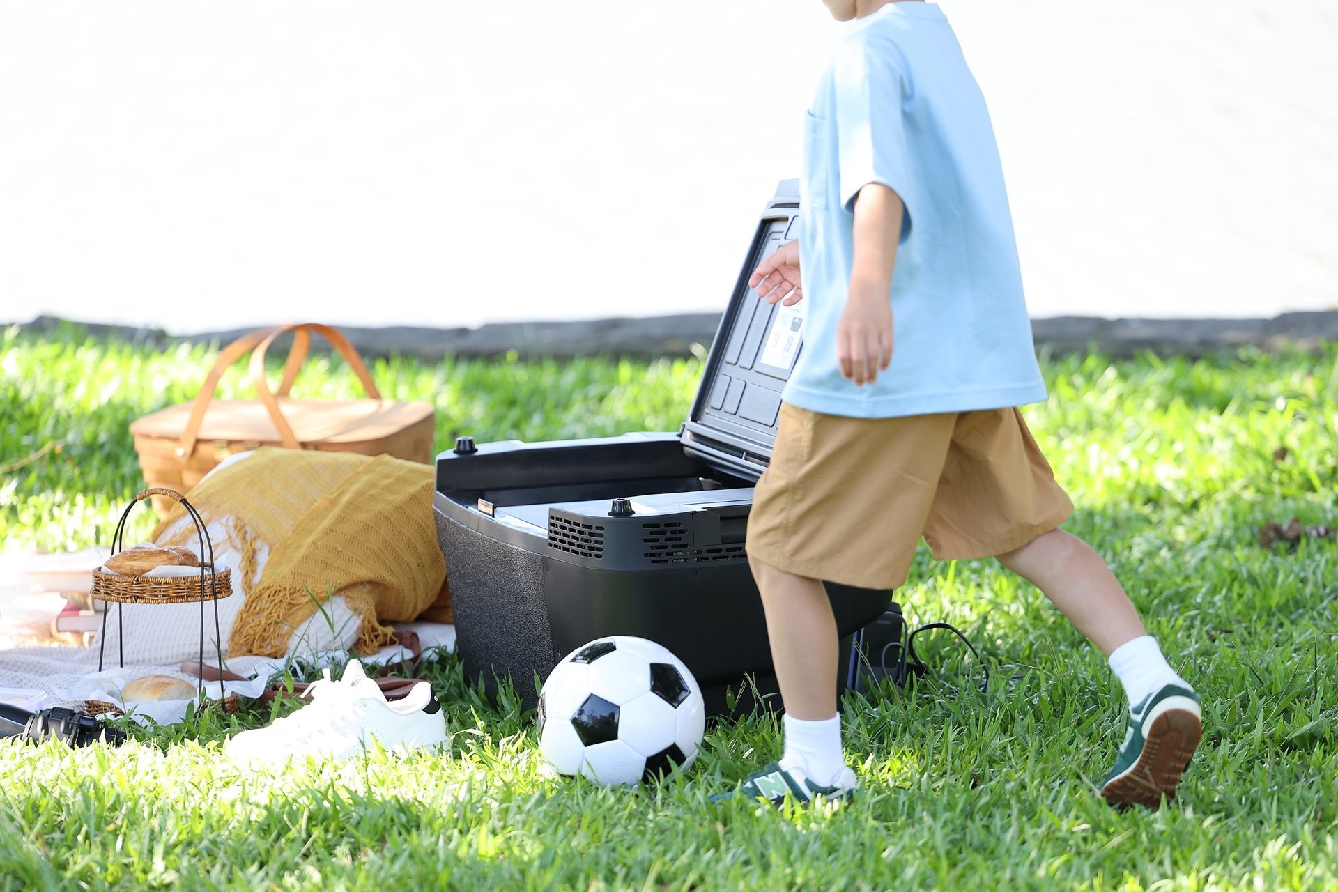 A child in shorts and a blue shirt walks near a picnic setup with baskets, shoes, a soccer ball, and the JOWUA Car Refrigerator for Tesla Model Y featuring intelligent dual-mode cooling on green grass. The bright sunlight illuminates the background.