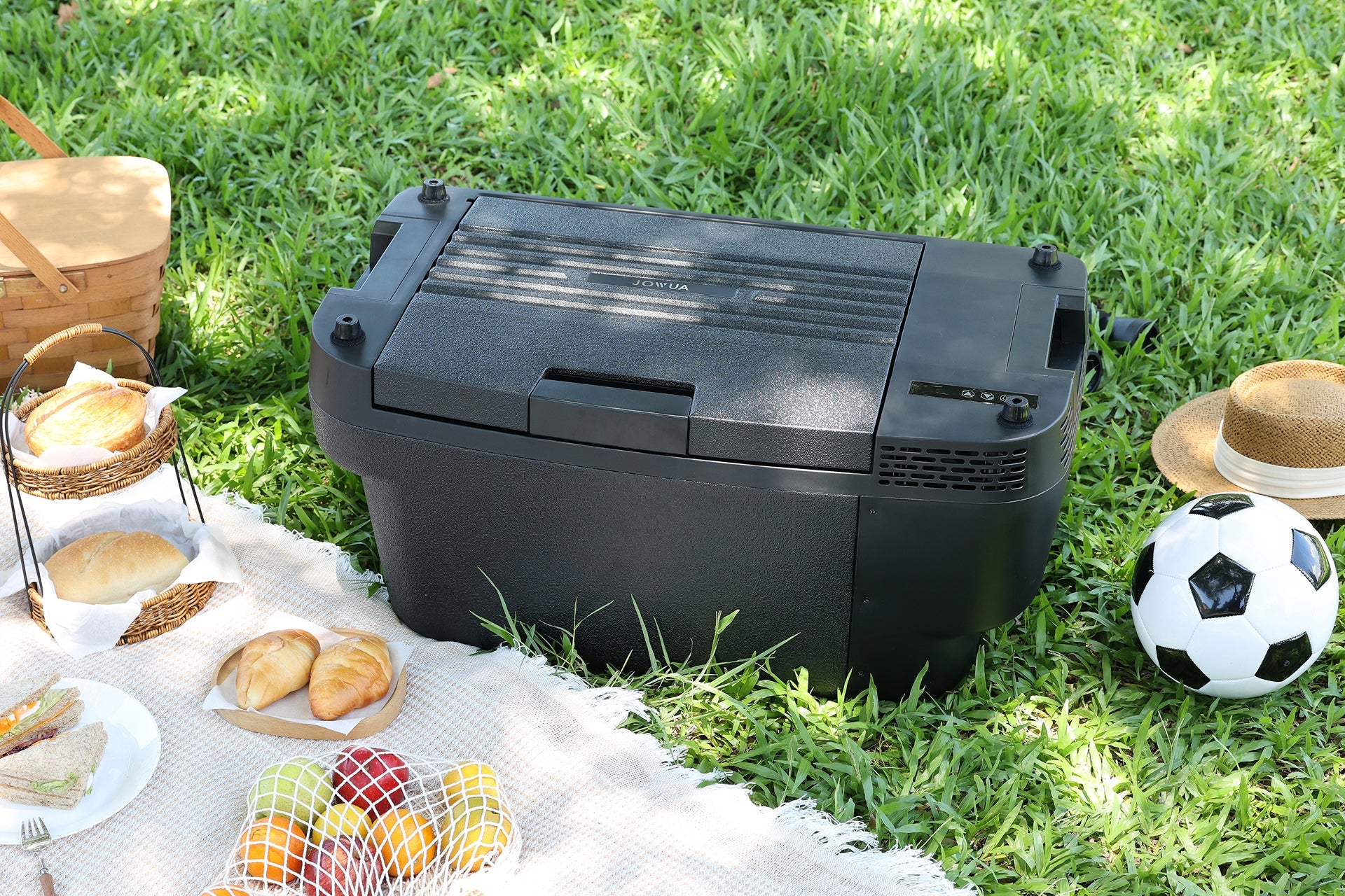 A JOWUA Car Refrigerator for Tesla Model Y, featuring intelligent dual-mode cooling, rests on green grass beside a picnic arrangement that includes bread, fruit, a basket, a soccer ball, and a straw hat.