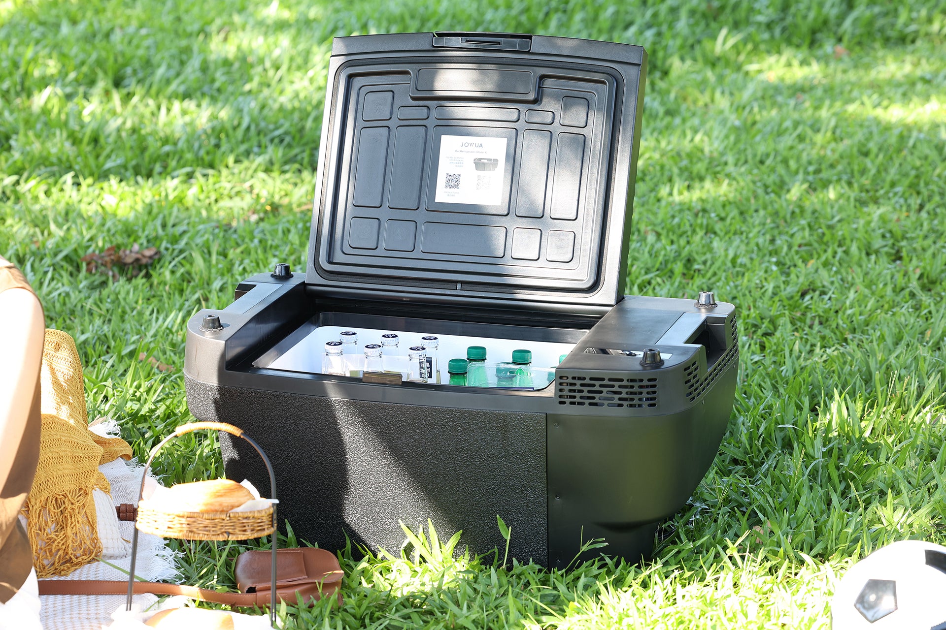 A JOWUA Car Refrigerator for Tesla Model Y, in sleek black, sits open on the grass, displaying canned and bottled drinks kept cold by its intelligent dual-mode cooling system. Powered conveniently via USB-C, this portable refrigerator ensures your beverages stay perfectly chilled. Nearby, a picnic basket and food hint at an enjoyable outdoor gathering.