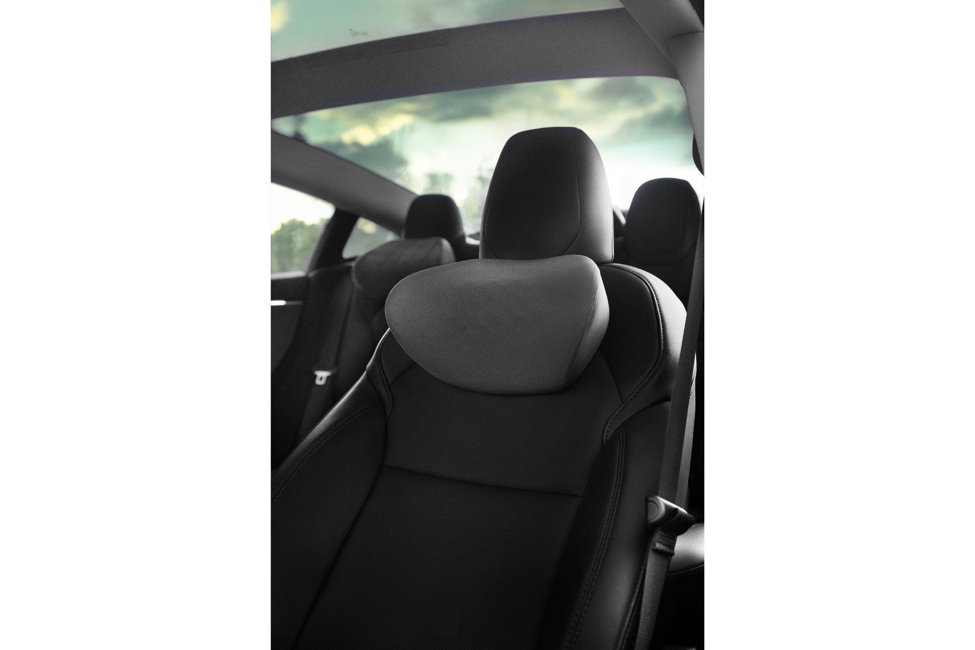 A close-up of a JOWUA Tesla Neck Pillow on a black leather car seat inside a vehicle, featuring the headrest and showcasing the panoramic glass roof with blurred outdoor scenery through the windows.
