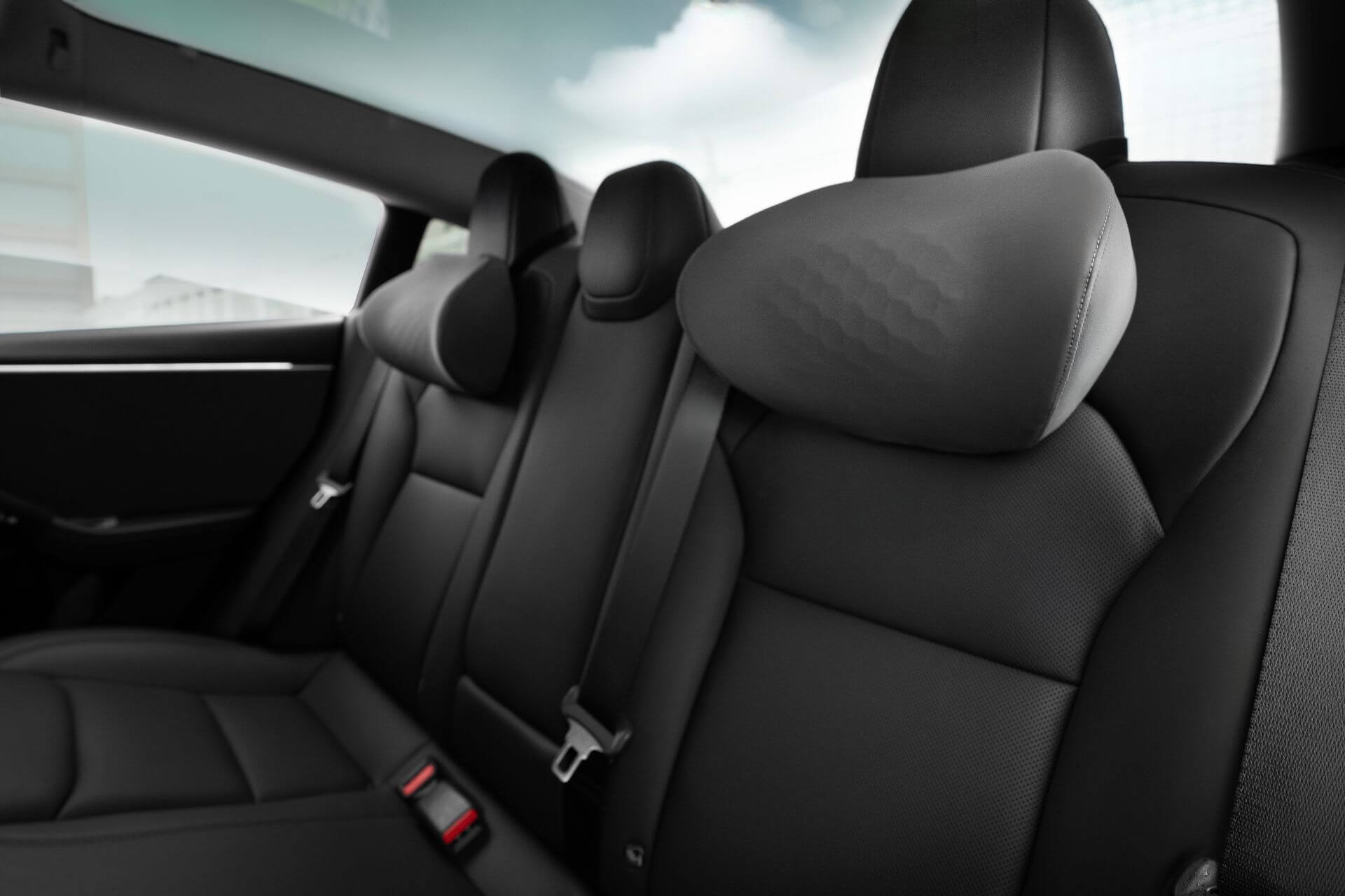 Close-up of a modern car’s back seat with three black leather ergonomic seats, headrests, seatbelt buckles, and a JOWUA Tesla Neck Pillow visible; a window shows a cloudy sky outside.