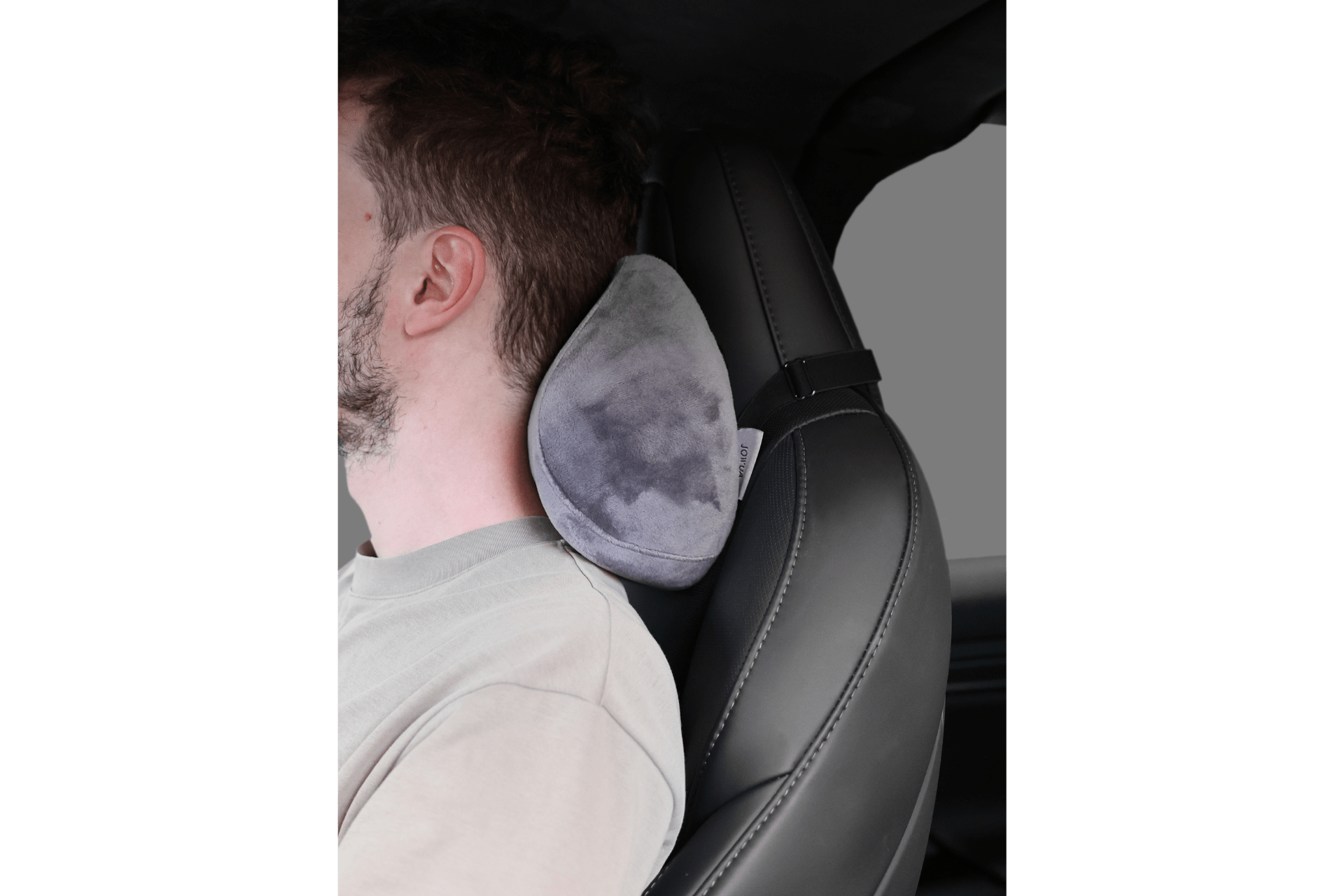 A person sits in a car seat, leaning back on the JOWUA Tesla Neck Pillow—a gray, plush memory foam pillow attached to the headrest—offering ergonomic support and comfort while seated.