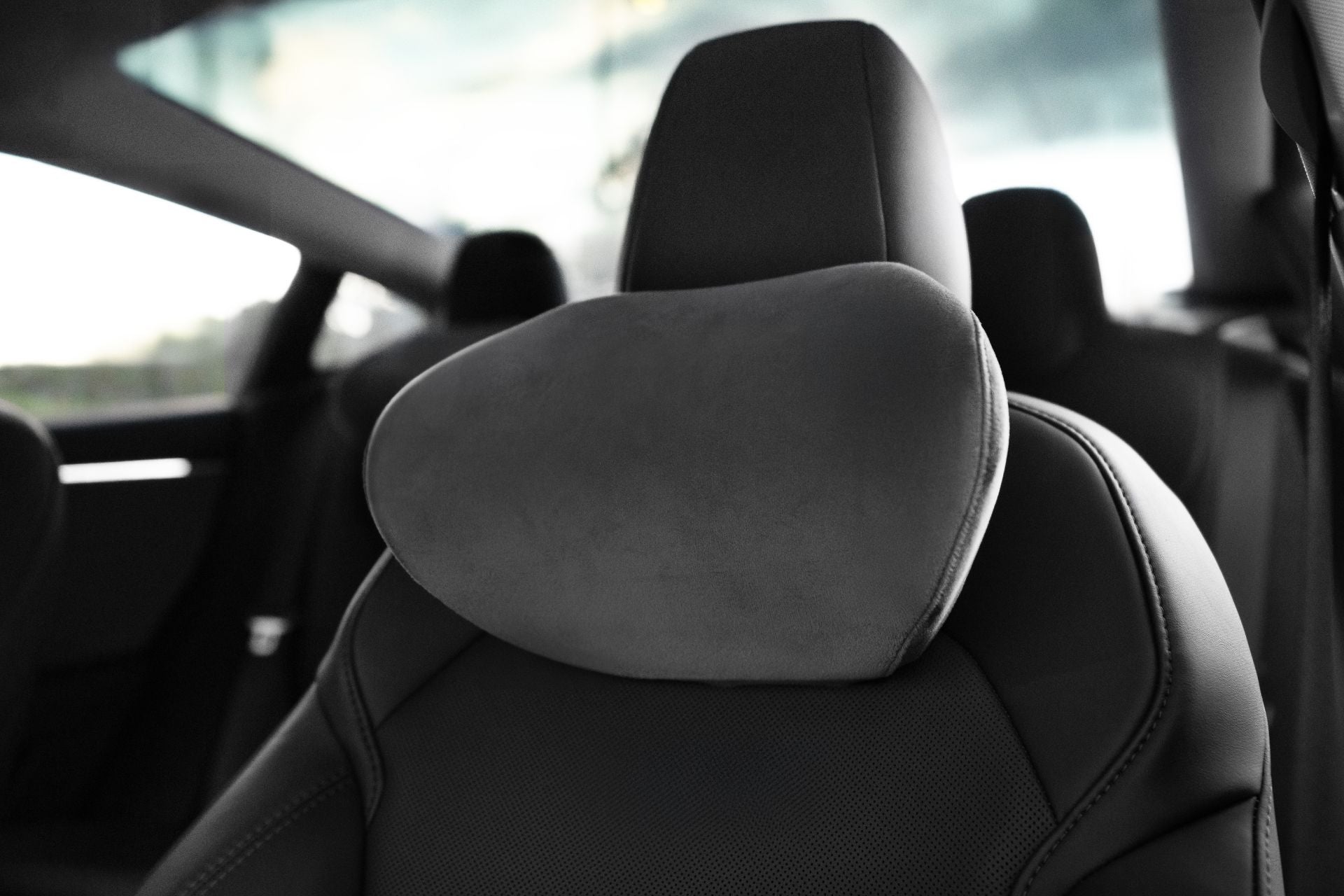 Close-up of a black car seat with the JOWUA Tesla Neck Pillow, a soft grey memory foam headrest, attached. The modern interior features ergonomic design, with blurred windows and extra seats in the background.