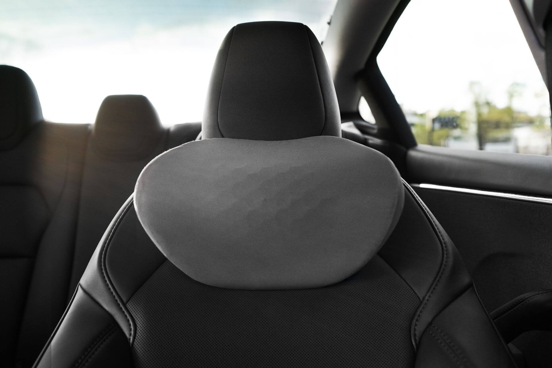 Close-up of a black car seat with a JOWUA Tesla Neck Pillow in gray memory foam attached, inside a modern vehicle. Sunlight streams through the windows onto the back seat in the background.
