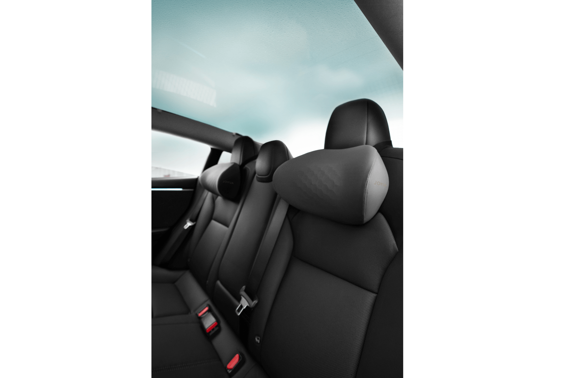 Backseat interior features JOWUA’s Tesla Neck Pillow on black leather seats with ergonomic headrests, seat belts, and red buckles; large rear window displays a cloudy sky outside.