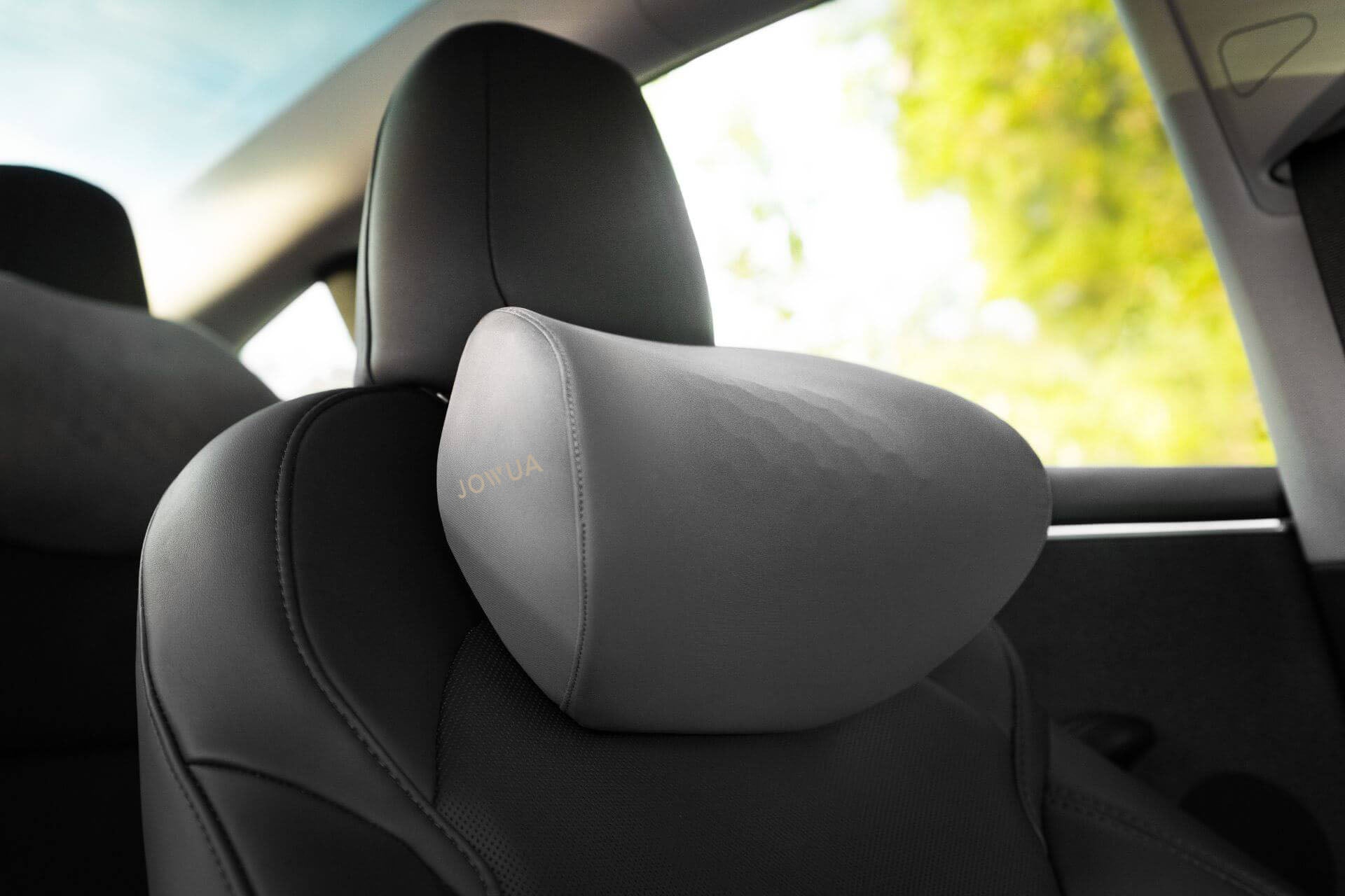 Close-up of a Tesla Neck Pillow by JOWUA, in gray memory foam with an ergonomic design, attached to a car seat headrest. Sunlight and green foliage are visible through the window in the background.