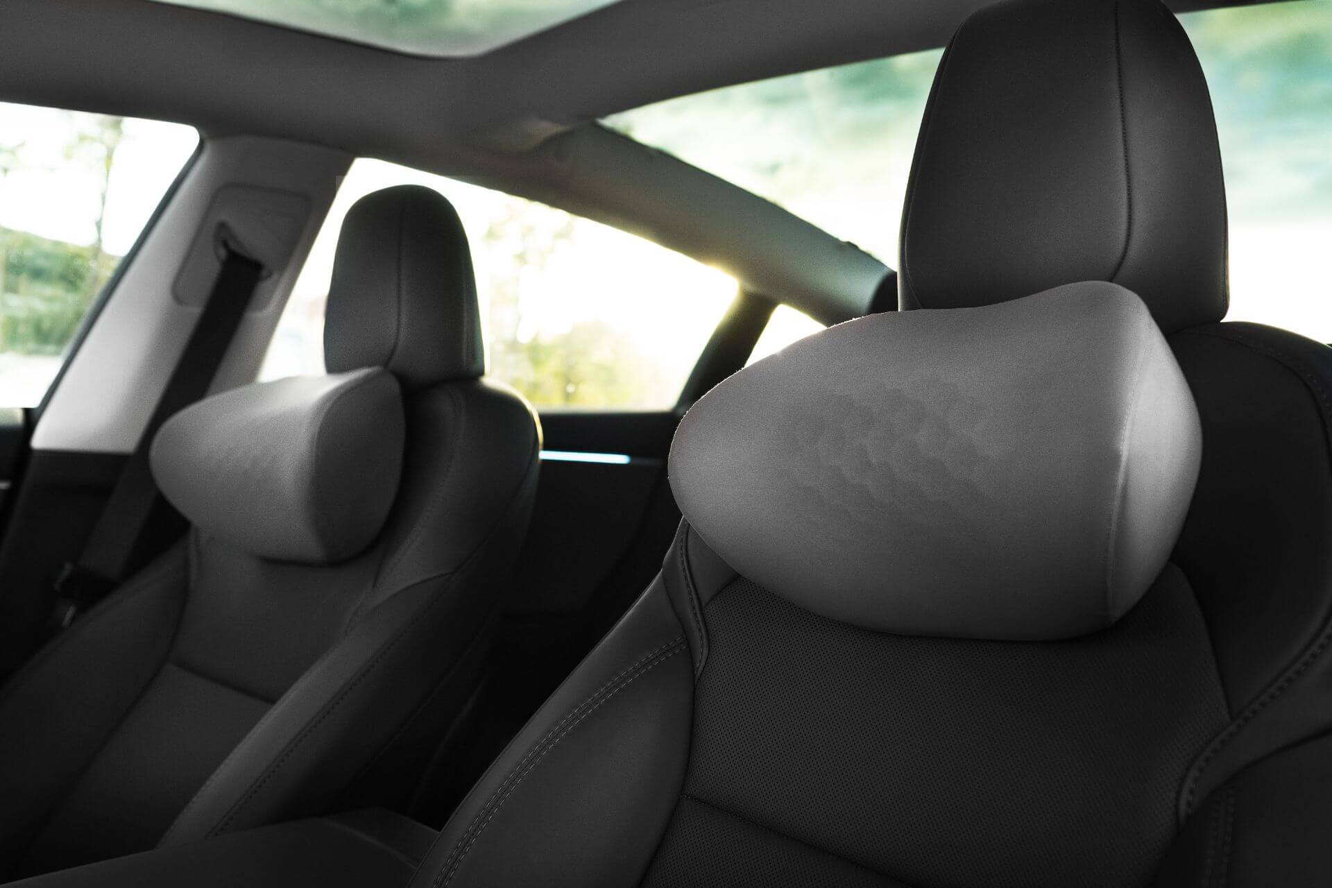 Close-up of a modern car’s front seats, featuring sleek black leather, ergonomic design with supportive headrests, and the JOWUA Tesla Neck Pillow for added comfort, all illuminated by natural light through side windows and a glass roof.