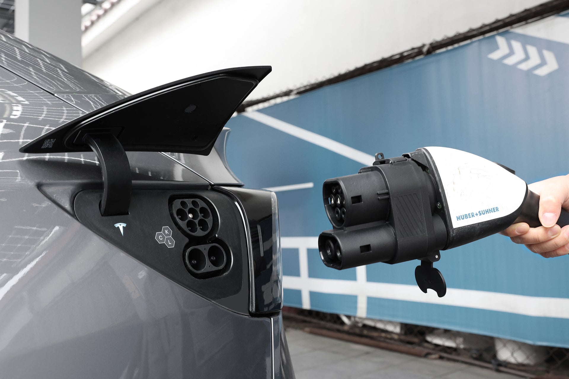 A person holds a JOWUA CCS1 to CCS2 DC & AC Combined Adapter near the open charging port of a grey Tesla, preparing to connect it. Blue and white walls with arrows appear in the background.