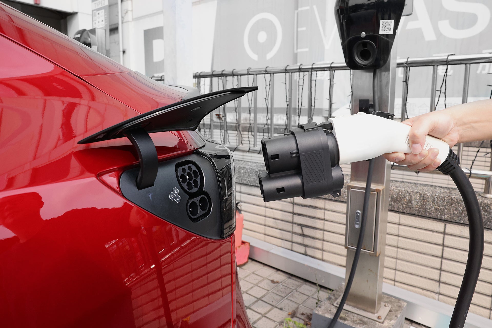 A hand holds the JOWUA CCS1 to CCS2 DC & AC Combined Adapter near the charging port of a red car, ready to connect it for fast charging at an EV station.