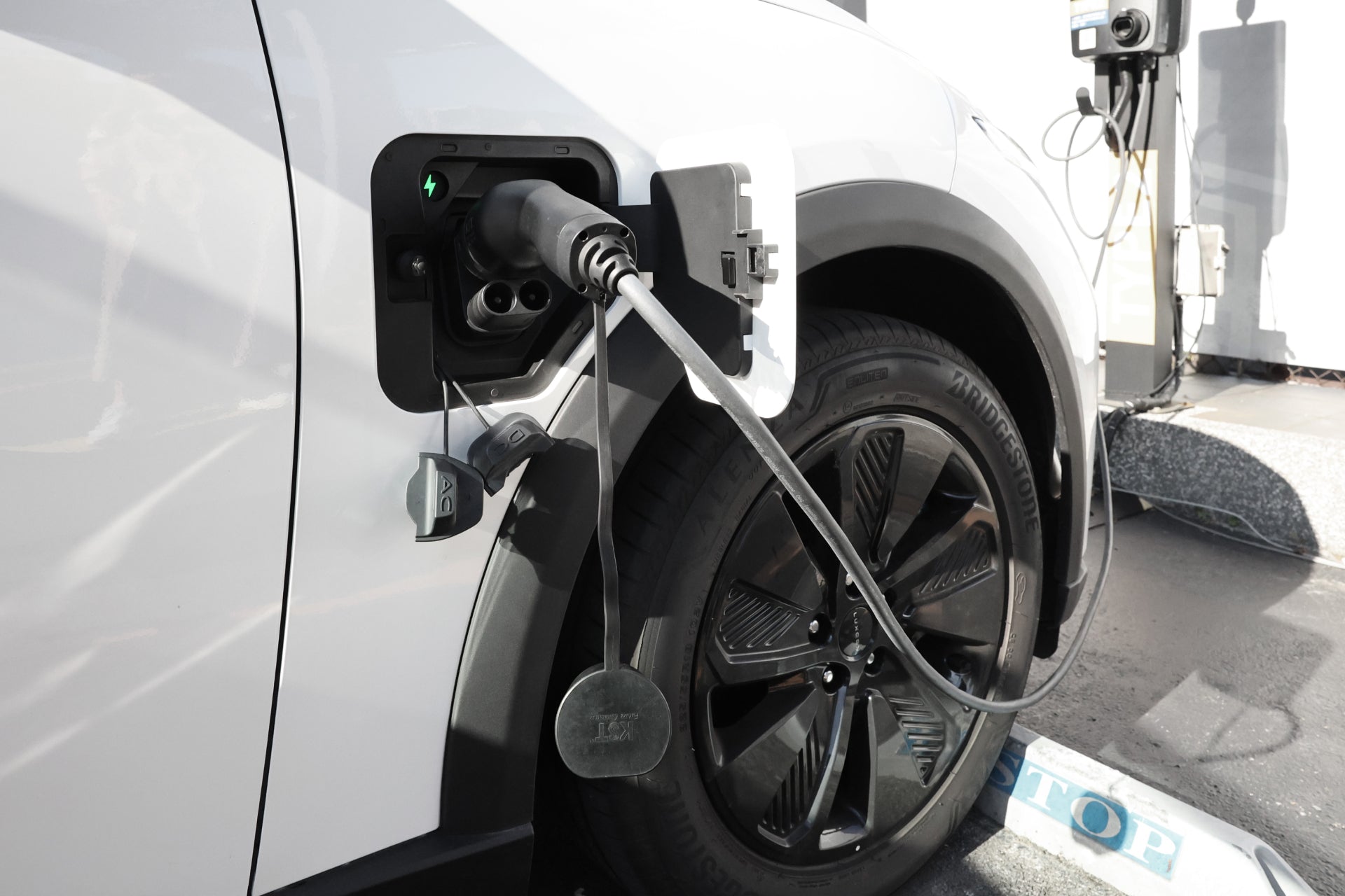 A white EV is charging at a fast station using the JOWUA CCS2 to CCS1 DC & AC Combined Adapter, with the cable plugged into the port near the front wheel and part of the tire visible.