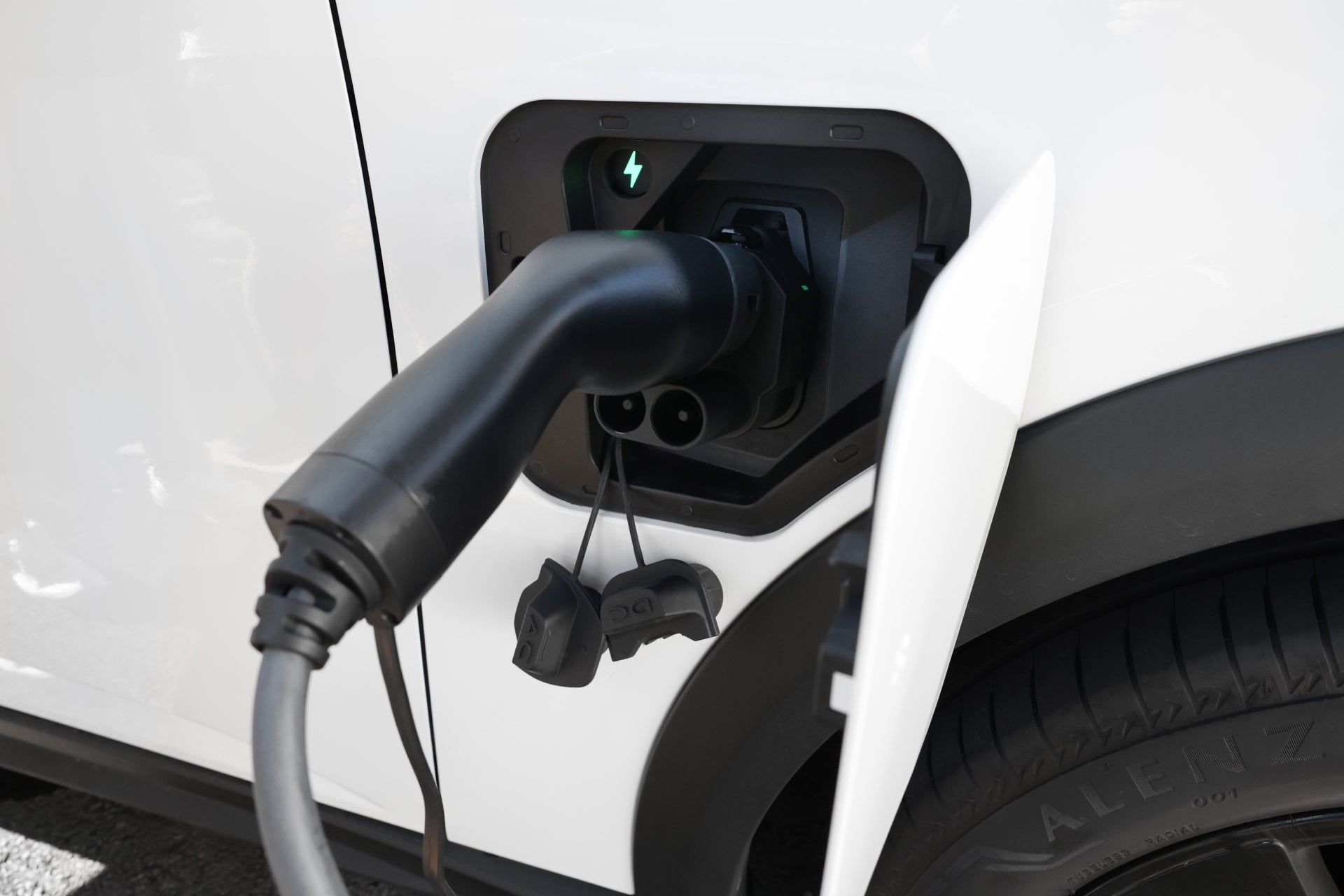 A close-up of a JOWUA CCS2 to CCS1 DC & AC Combined Adapter charging an electric car, with the cable connected to the side port, indicator lights on, and part of the rear tire in view.