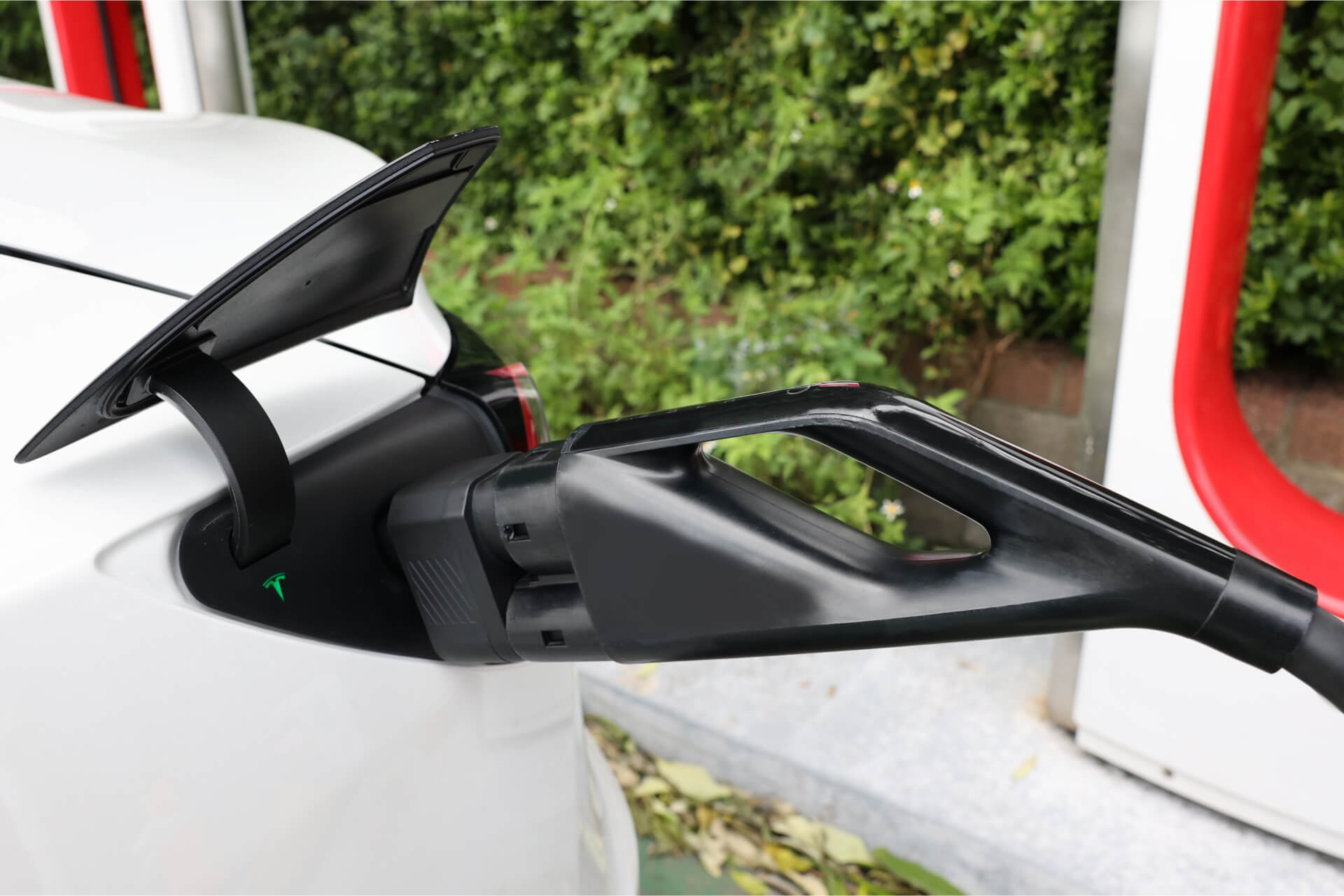 A close-up of a white electric car charging with the JOWUA CCS2 to NACS DC Charging Adapter, with the black adapter plugged in and greenery softly blurred in the background. The image highlights the vehicle's Tesla CCS compatibility.
