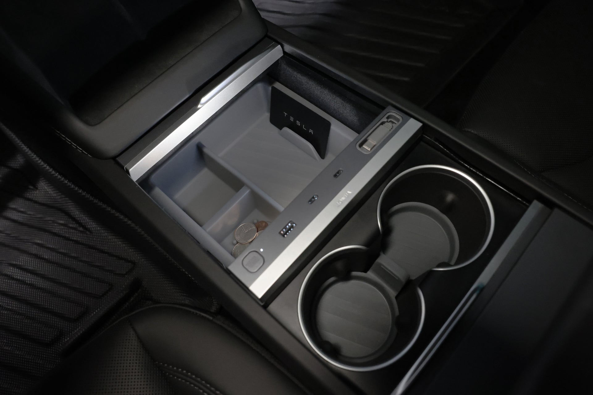 In the car's sleek and modern interior, the LED Center Console Combo by JOWUA for the Tesla Reengineered Model 3 Highland (2024+) features an open center console system with a storage tray designed to hold coins and a USB port, alongside two circular cup holders. The design is enhanced by black upholstery and includes a silicone organizer to keep essentials tidy and accessible.