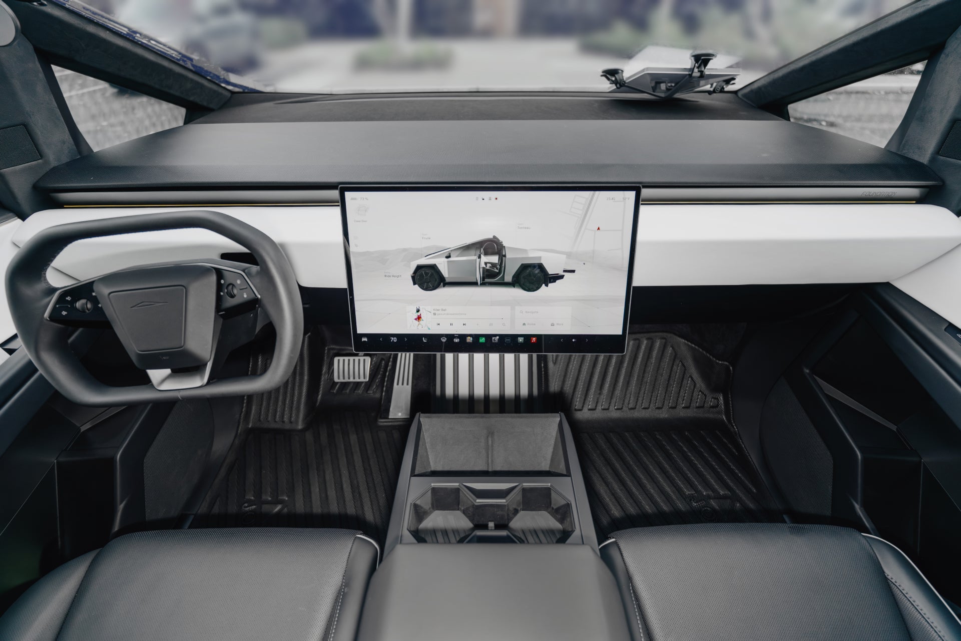 A futuristic vehicle interior with black seats and JOWUA All Weather Floor Liners (Tesla Cybertruck) featuring geometric patterns and raised edge coverage, a minimalist dashboard, rectangular steering wheel, and large touchscreen displaying a truck image.
