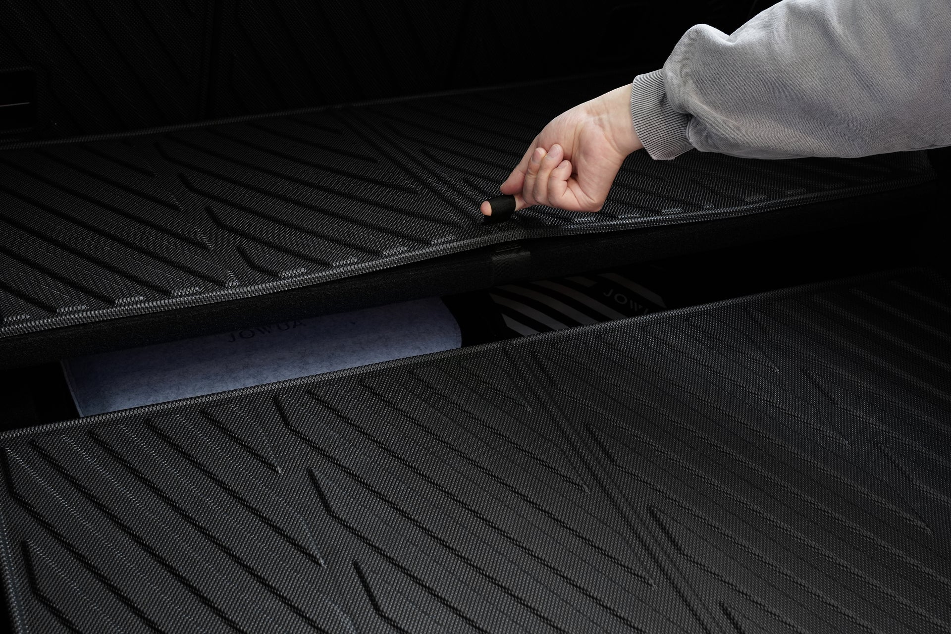 A hand lifts the textured black JOWUA Model Y EasyShield Waterproof Trunk Liner + Rear Seats Back Cover Bundle (Juniper 2025+/2018-2024), revealing extra storage space beneath.