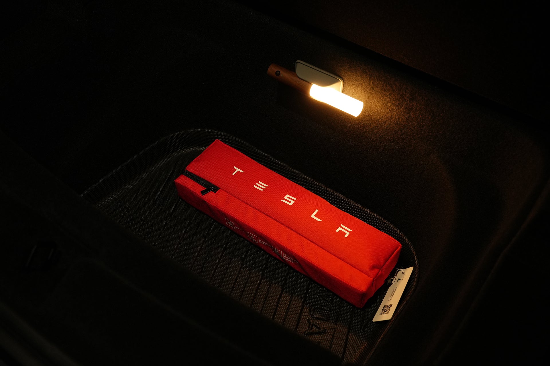 A red Tesla-branded pouch rests on a black JOWUA Model Y EasyShield Waterproof Trunk Liner + Rear Seats Back Cover Bundle in the dimly lit trunk of a Tesla Model Y, softly illuminated by an overhead interior light.