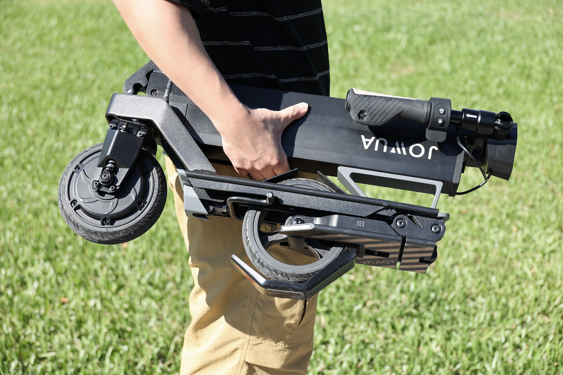 A person in beige pants and a dark striped shirt carries a folded black JOWUA Folding Electric Scooter|Performance outdoors on grass.