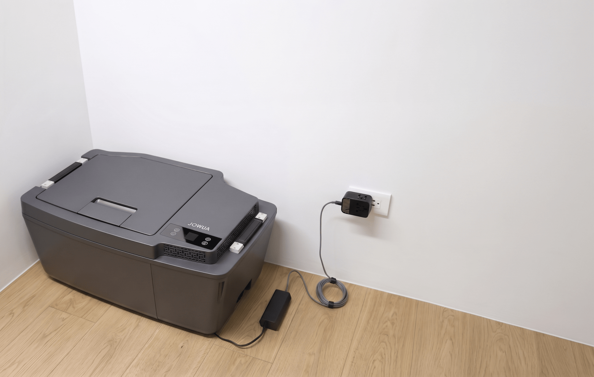 A JOWUA Car Refrigerator for Tesla Model 3 (Premium/Performance 2024-2026+) sits on a light wood floor near a white wall, plugged in with a USB-C power system. The area around the refrigerator and wall is uncluttered.