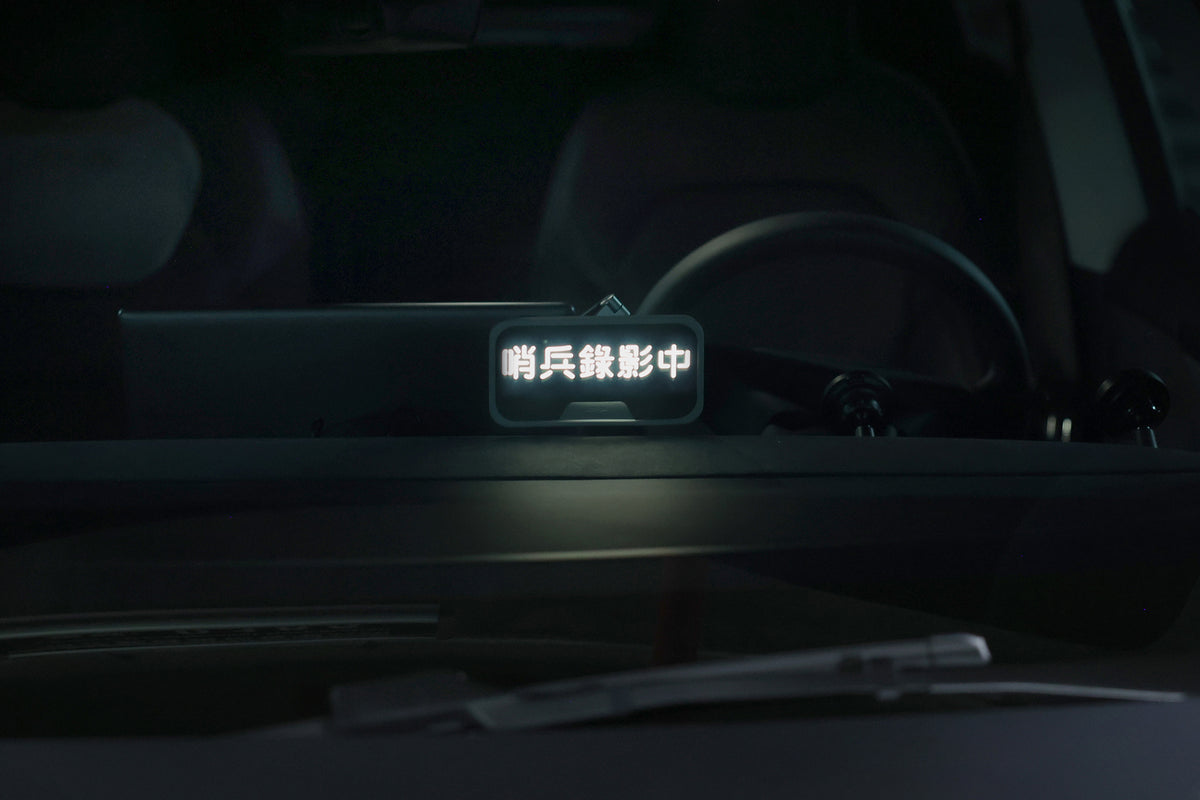A close-up of a car dashboard features a small illuminated display with Chinese characters in white, viewed through the windshield. The dimly lit interior reveals a partially visible steering wheel, while the JOWUA MagSafe LED Light with Light Stencil Set casts a subtle glow that complements the sleek setup.