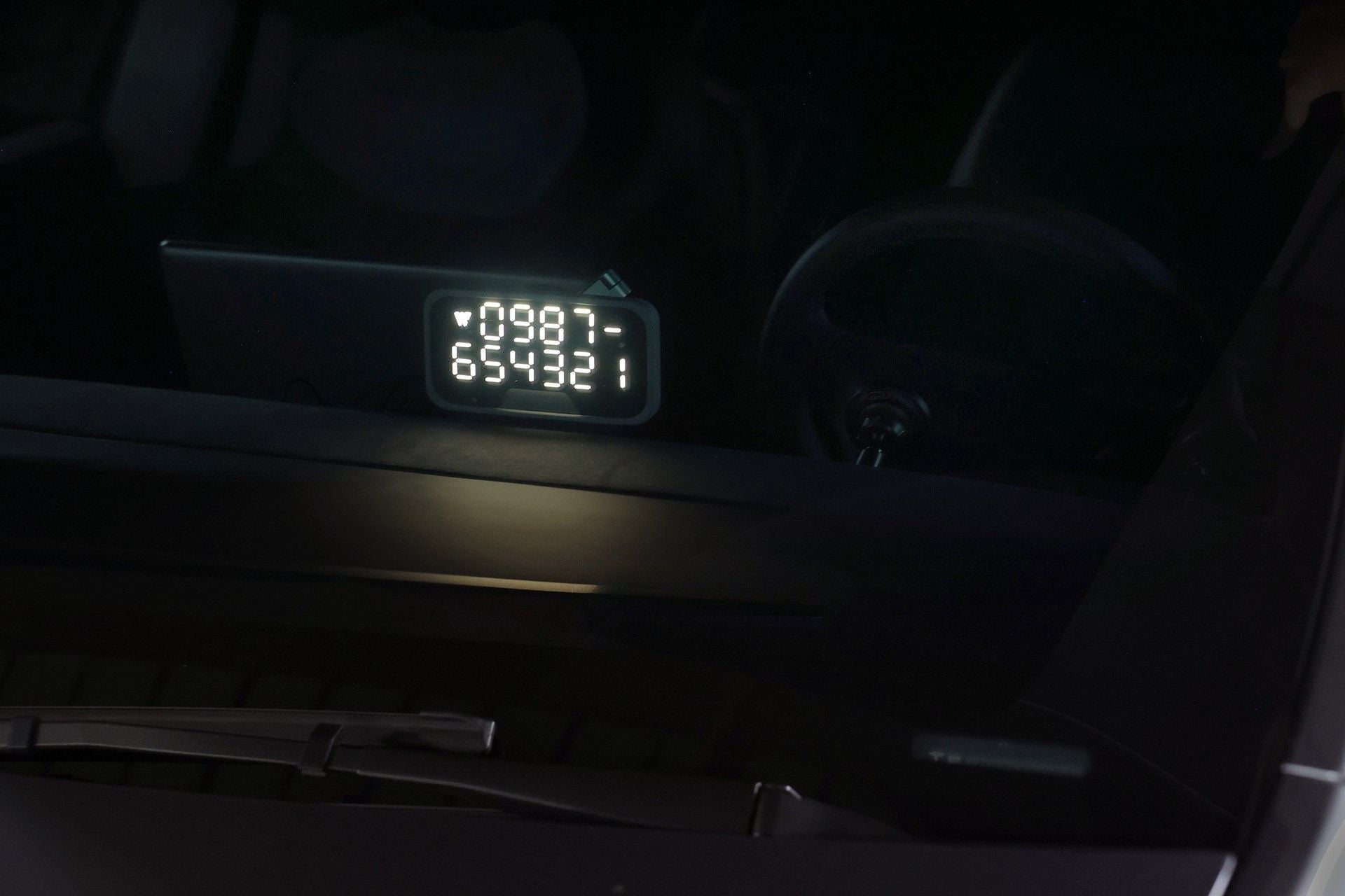 A digital display shows the numbers 0987 and 654321, seen through a car windshield with part of the dashboard and steering wheel visible in low light. On the console, a JOWUA MagSafe LED Light with Light Stencil Set adds subtle illumination, emphasizing seamless wireless charging and customizable lighting effects for your devices even on nighttime drives.