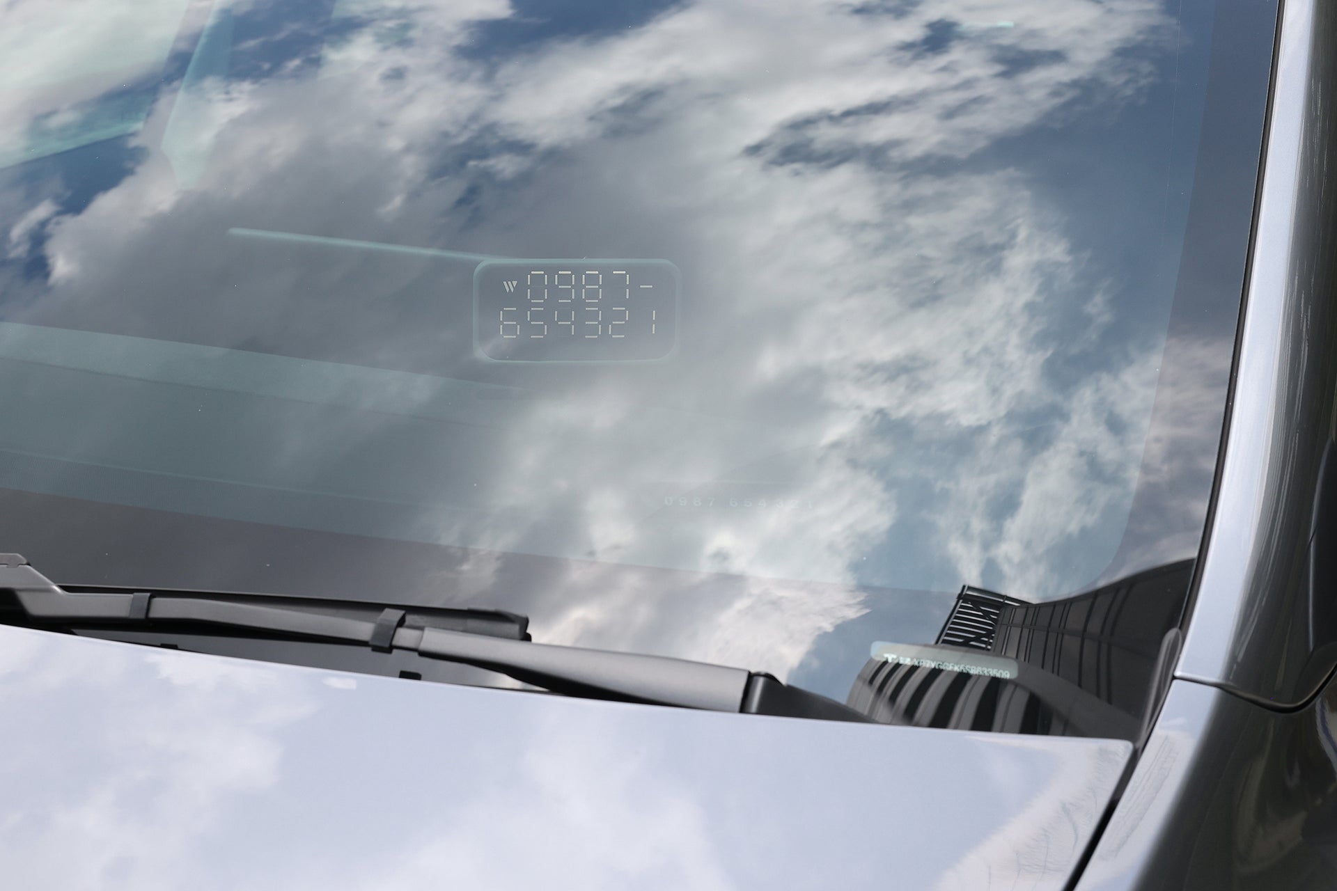 A car windshield reflects a cloudy sky. Inside the windshield, a digital display shows numbers and letters—likely a heads-up display—while a wiper rests at the bottom. On the dashboard, the JOWUA MagSafe LED Light with Light Stencil Set casts a subtle glow beside a Tesla wireless charging pad, blending modern convenience with sleek design.
