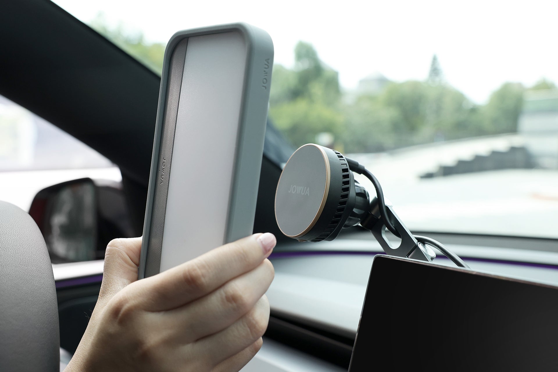 A person holds the JOWUA MagSafe LED Light with Light Stencil Set—a rectangular device—near a circular car phone mount attached to a car dashboard, with a blurred outdoor scene visible through the window. The setup features convenient wireless charging, ensuring reliable power delivery while on the go.
