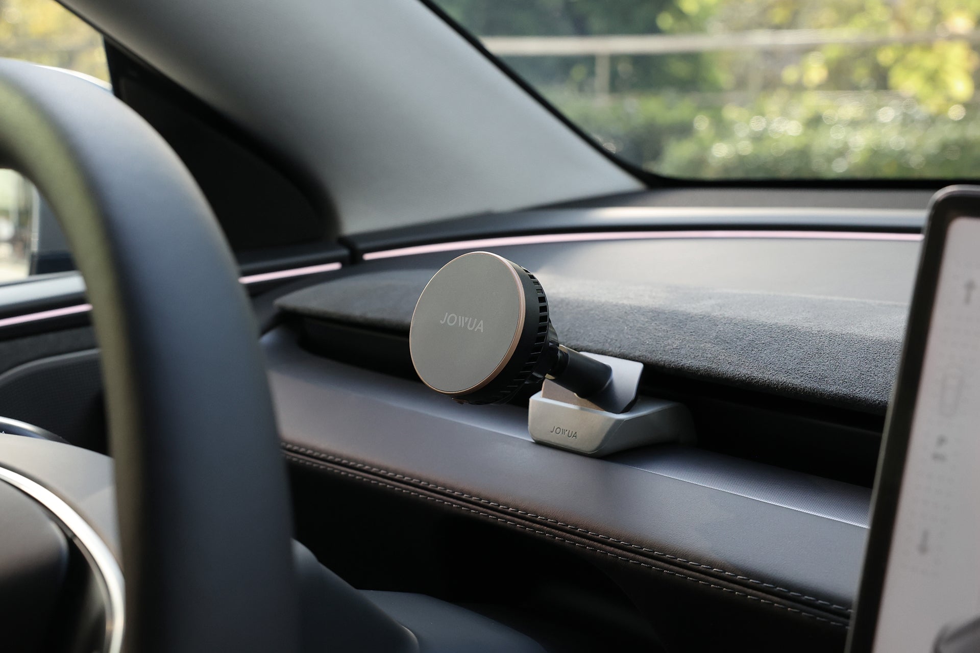 A close-up view of a JOWUA Active Cooling MagSafe Car Mount for Air Vents (Model 3 Highland & Model Y Juniper) attached inside a car, with a blurred green outdoor background visible through the windshield.