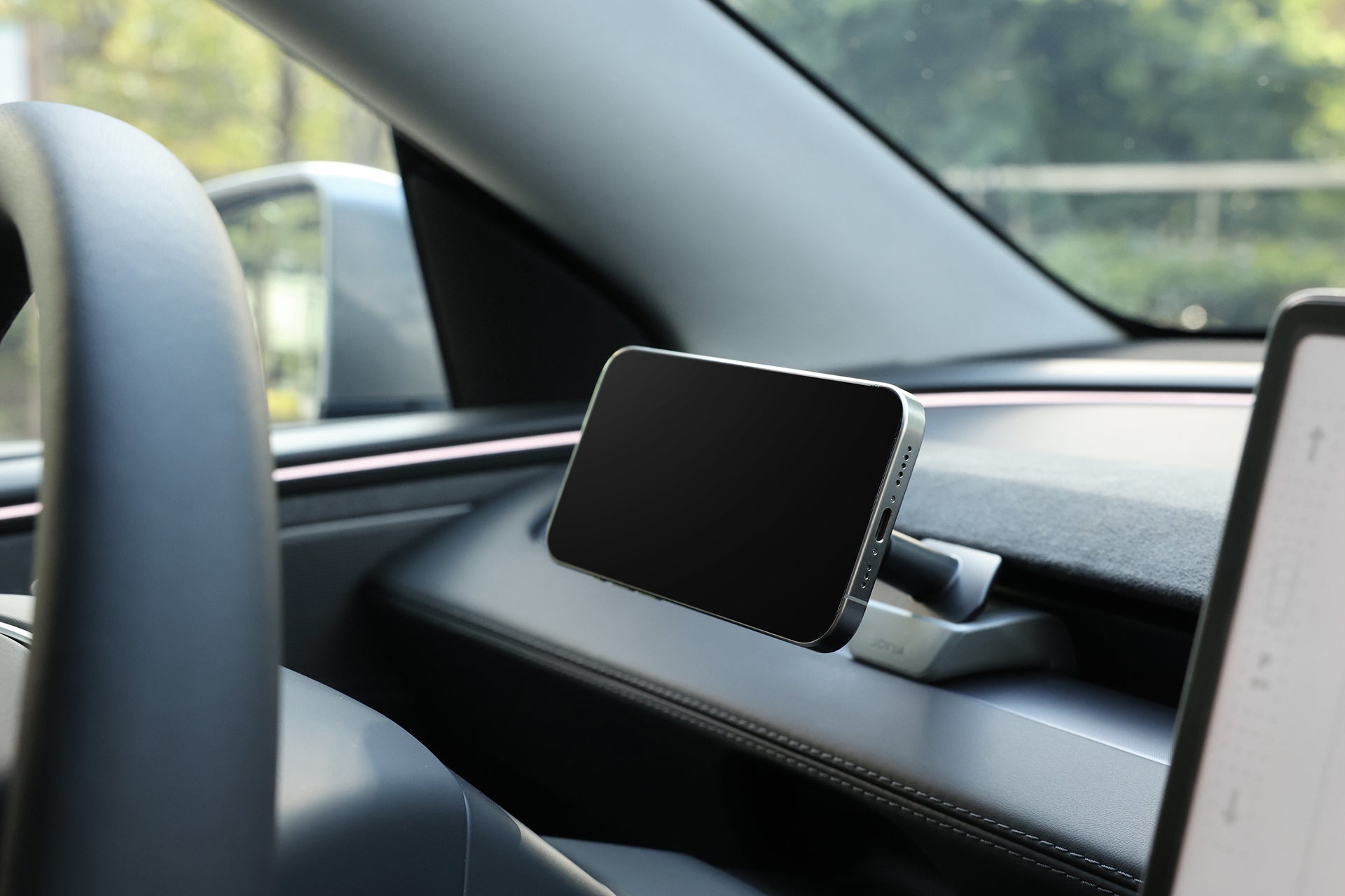 A smartphone mounted horizontally on the JOWUA Active Cooling MagSafe Car Mount for Air Vents (Model 3 Highland & Model Y Juniper), inside a modern car with part of the steering wheel, display, and sunlit trees visible through the window.