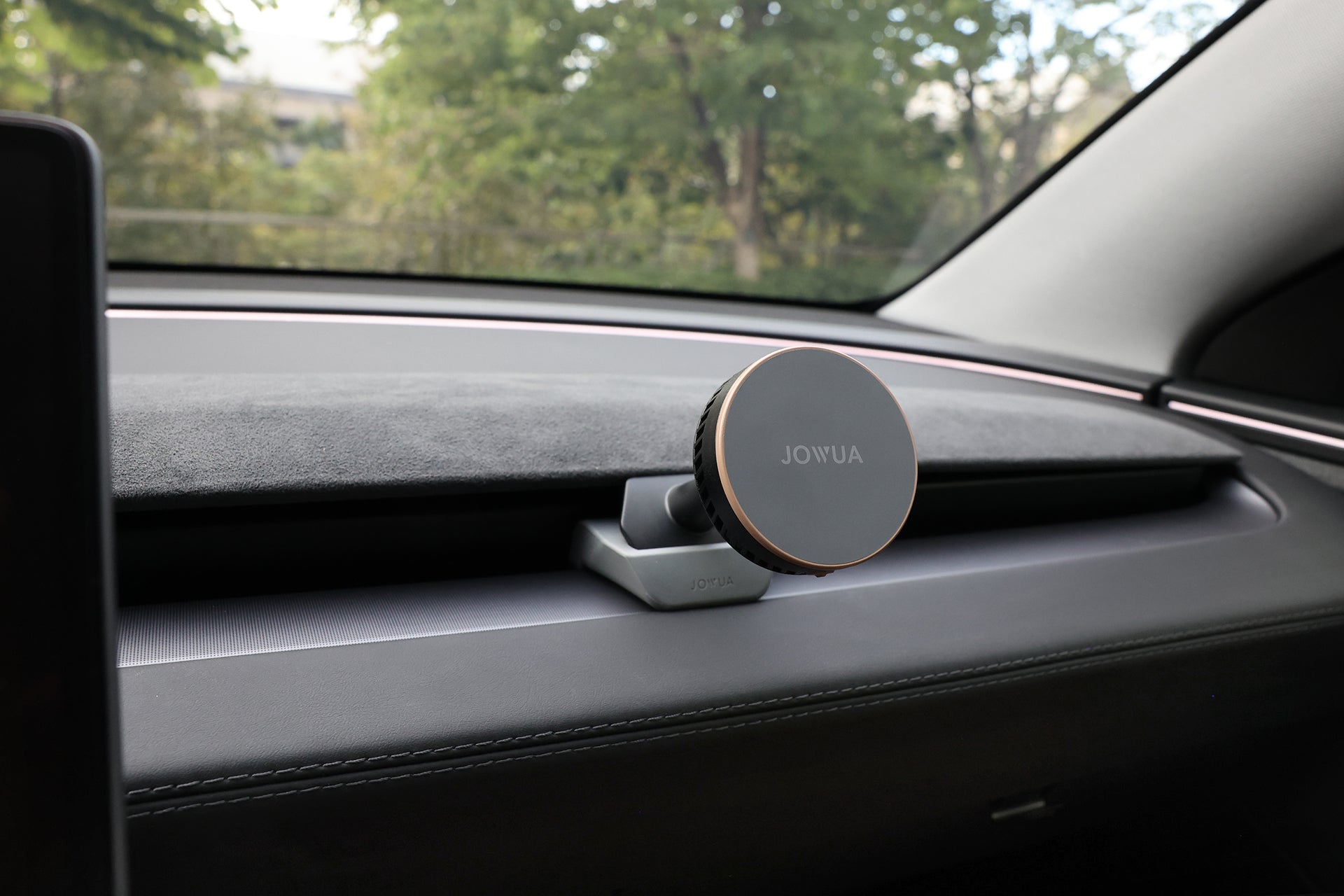 A JOWUA Active Cooling MagSafe Car Mount for Air Vents (Model 3 Highland & Model Y Juniper) is attached to a car dashboard, providing wireless charging, with trees visible through the window.