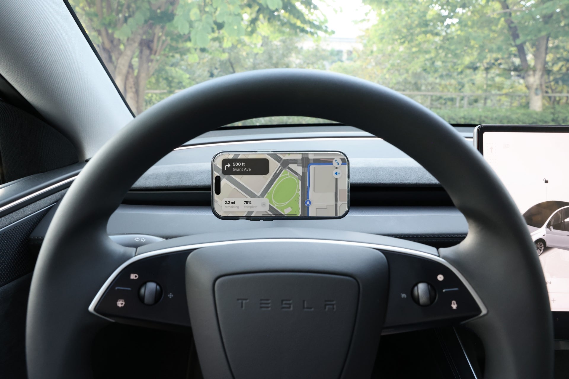 A JOWUA Active Cooling MagSafe Car Mount for Air Vents (Model 3 Highland & Model Y Juniper) holds a smartphone above a Tesla steering wheel, displaying navigation, with trees and greenery visible through the windshield.