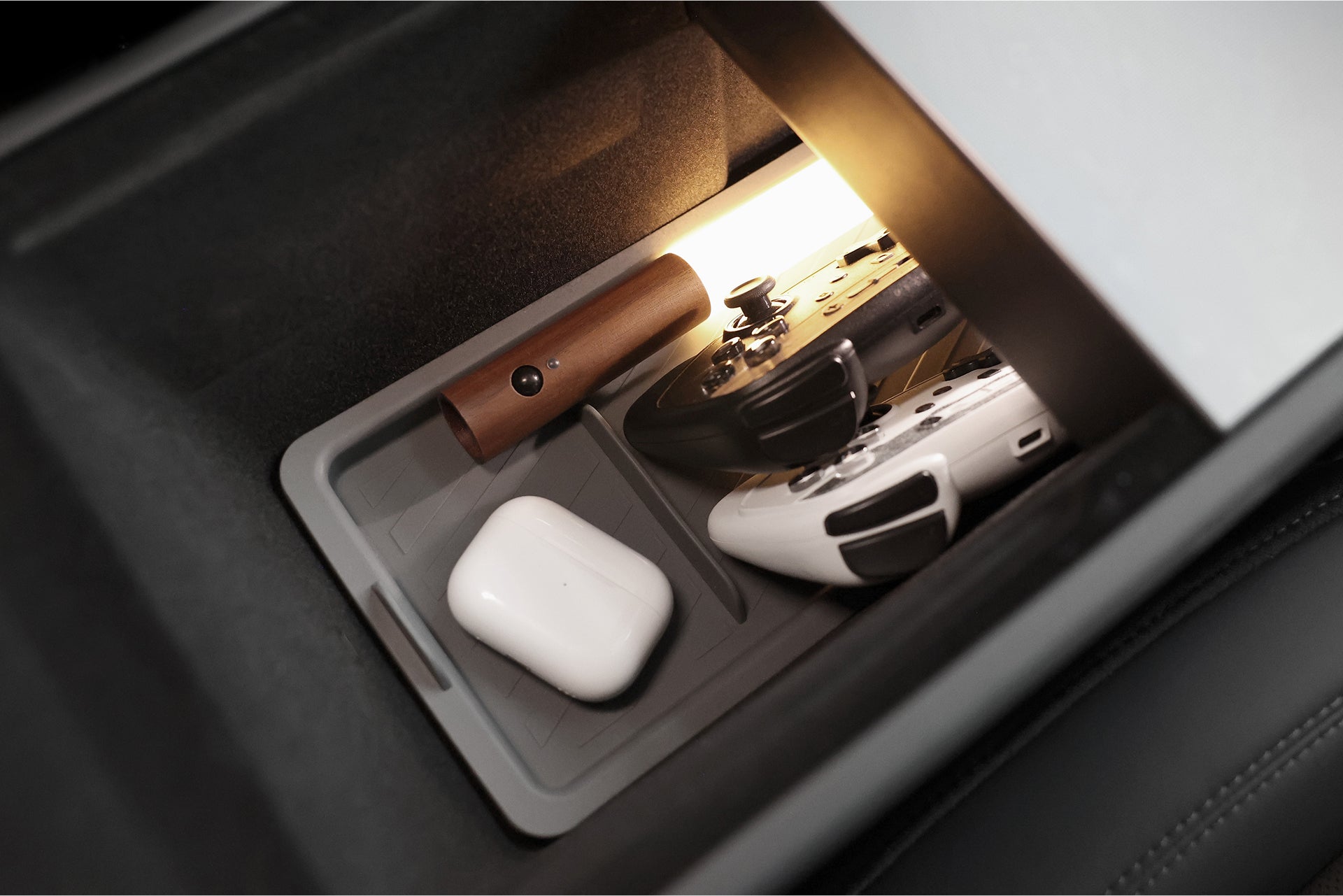 The JOWUA Armrest Organizer Set for Tesla Model Y Juniper 2025+/Model 3 Highland 2024+ neatly holds wireless earbuds in a white case, a small brown cylinder, and two game controllers, all illuminated by its built-in light.