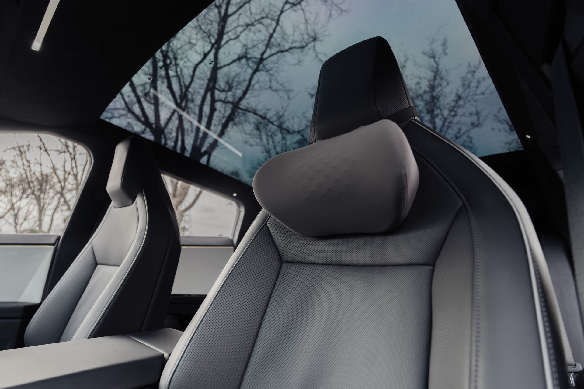 Close-up of a JOWUA Tesla Neck Pillow on a modern black car seat under a large glass roof, with tree branches and sky outside; focus is on the front seat’s headrest.