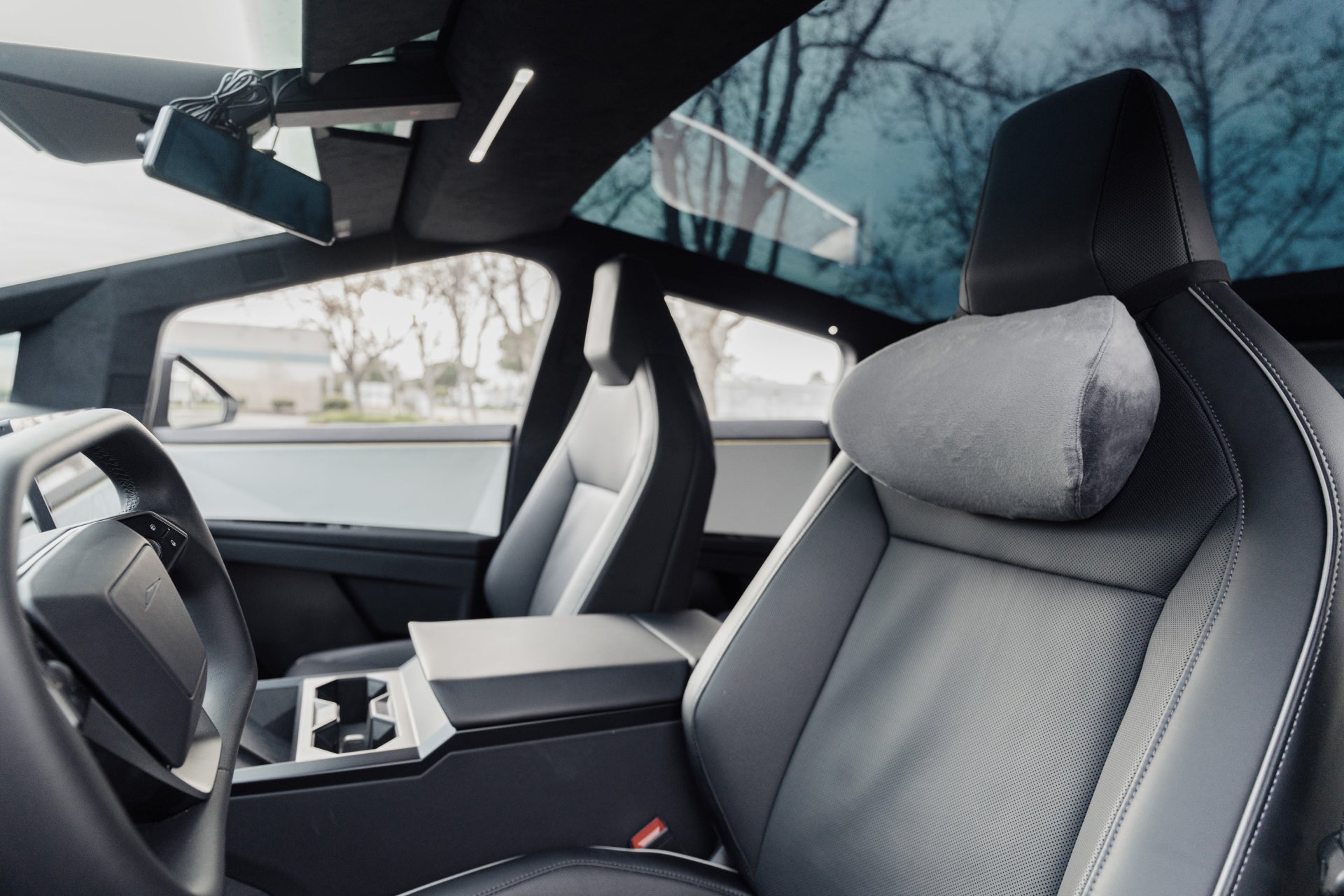 Close-up of the front seats and dashboard inside a modern car, featuring a JOWUA Tesla Neck Pillow, black leather upholstery, ergonomic design, and a large glass roof with tree branches visible outside.