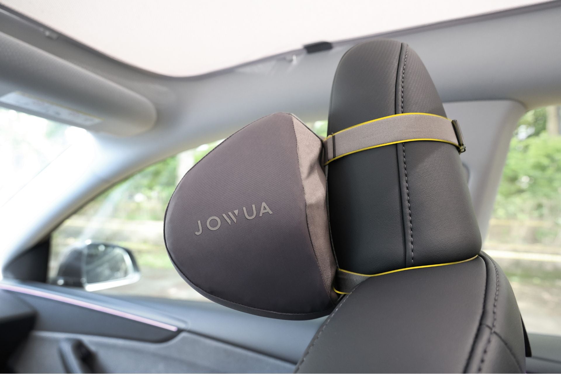 A gray JOWUA Tesla Neck Pillow with ergonomic design is attached to a car seat headrest using yellow elastic straps, inside a car with a light-colored ceiling and green trees visible through the window.
