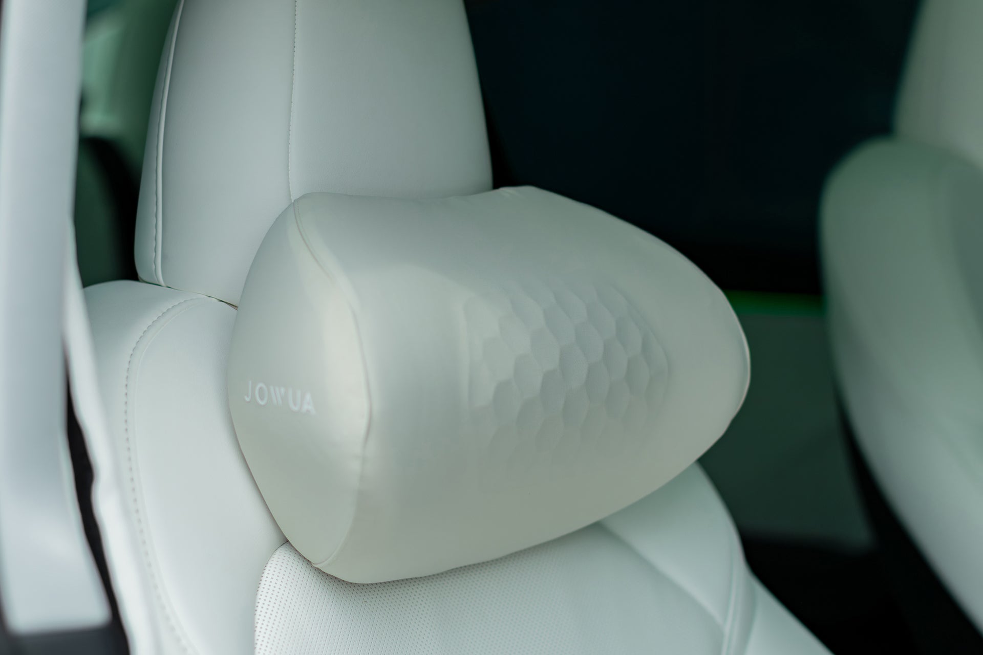 Close-up of a Tesla Neck Pillow by JOWUA, attached to a white car seat headrest. With an ergonomic honeycomb design and smooth leather-like texture, the neck cushion features the JOWUA brand name.