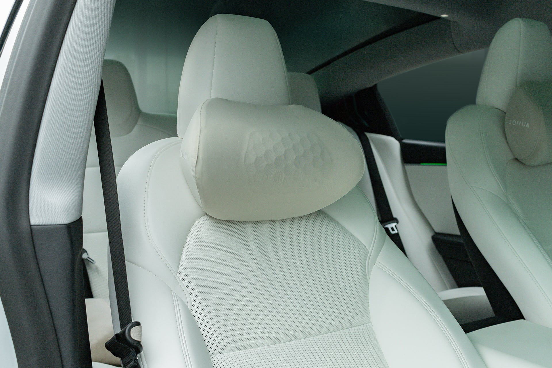 Close-up of a modern car's front passenger seat featuring white leather upholstery and a JOWUA Car Neck Pillow attached to the headrest. The back seat and seat belt are visible in the background.