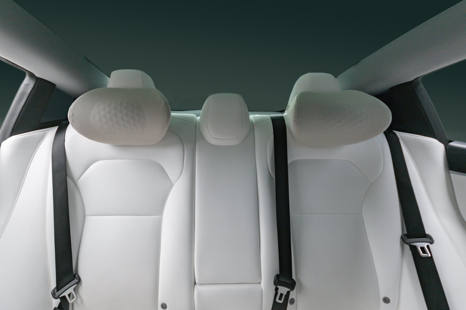 The image displays the backseat of a car featuring three white leather seats and JOWUA Tesla Neck Pillows with ergonomic, padded, and curved headrests. Two black seat belts and a dark window complete the clean, modern look.