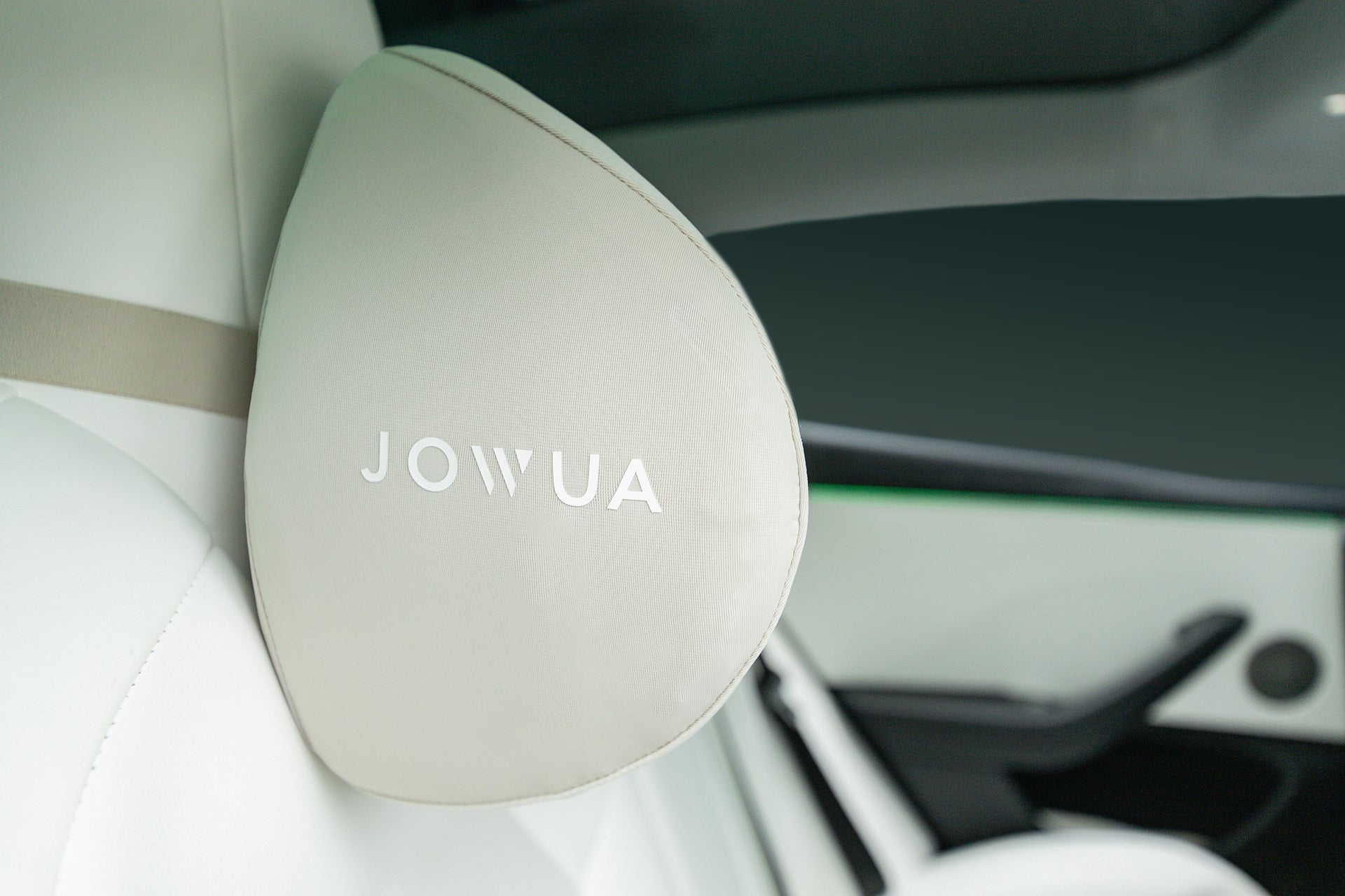 A close-up of the JOWUA Tesla Neck Pillow in light gray memory foam with "JOWUA" printed on it, showcasing its ergonomic design as it attaches to the seat of a modern car with a white interior.