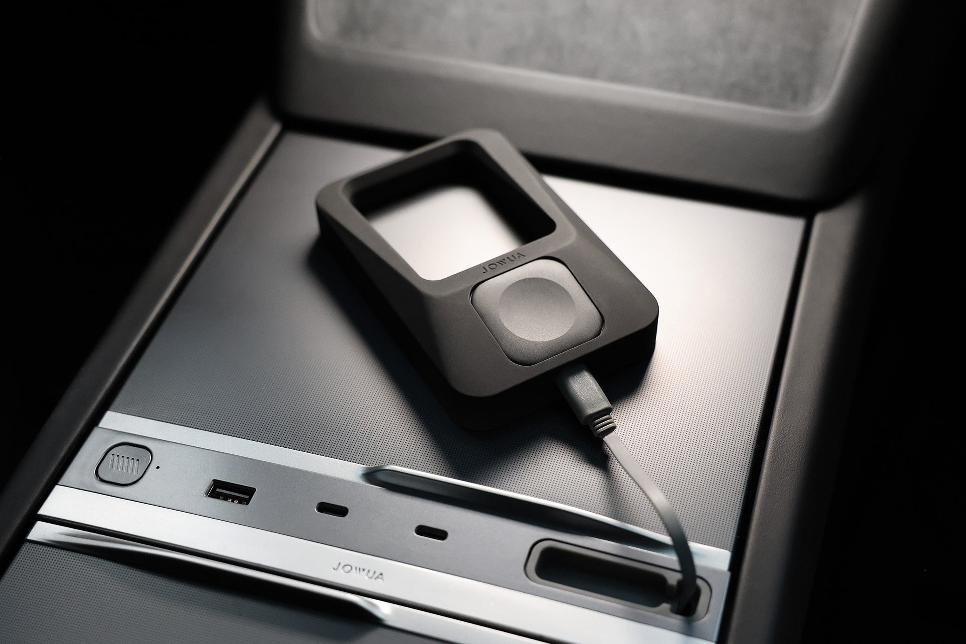The JOWUA Tesla Everyday Essentials MagSafe car mount, featuring a black rectangular open frame and circular button, sits on a metallic center console with a USB-C port—ideal for seamless Apple ecosystem charging.