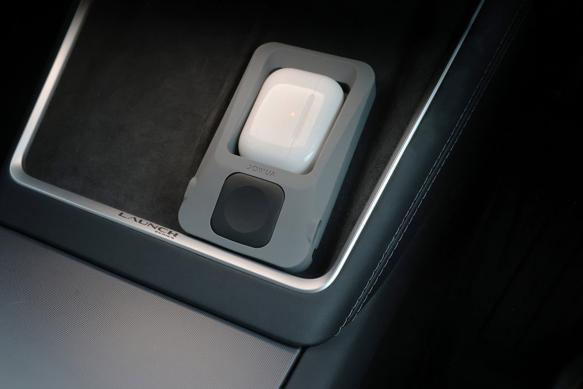 A pair of wireless earbuds in a gray charging case sits in a Tesla’s center console storage area, next to the JOWUA Power Dock Combo accessory charger with “LAUNCH” visible on the trim.