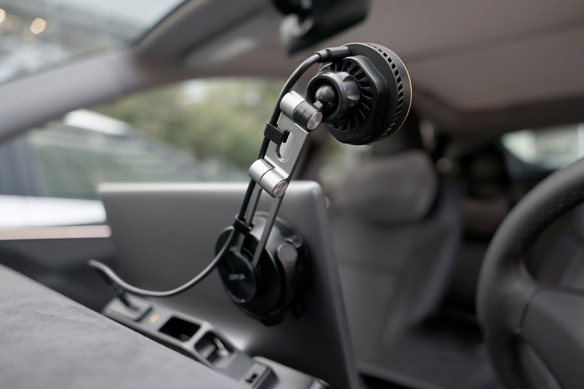 A close-up of a professional microphone mounted inside a car, attached to the dashboard near the JOWUA Active Cooling Tesla Compatible Phone Mount and connected by cable, with blurred steering wheel and seats in the background.