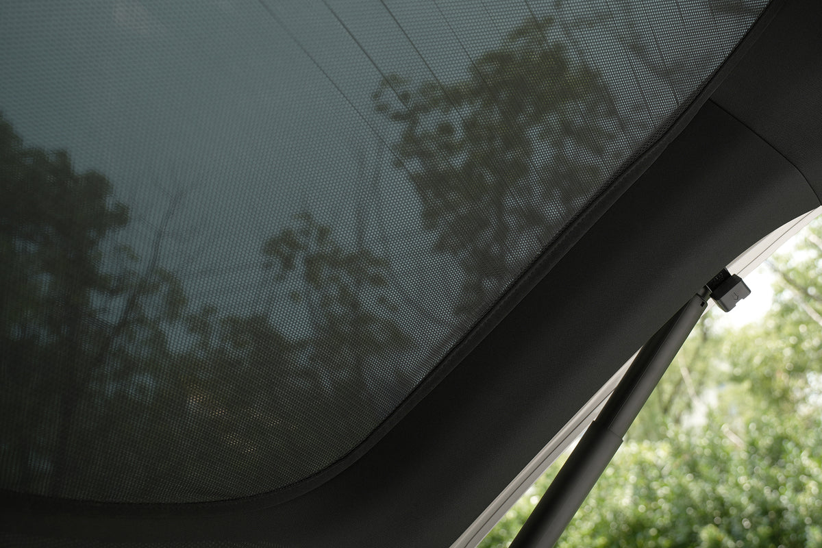 A view from inside a car highlights the JOWUA Rear Lift Gate + Triangular Window Sunshade for Model Y L (2026+), custom-fitted on the window, with blurred trees and power lines visible outside, creating a shaded and partially obscured outdoor scene.