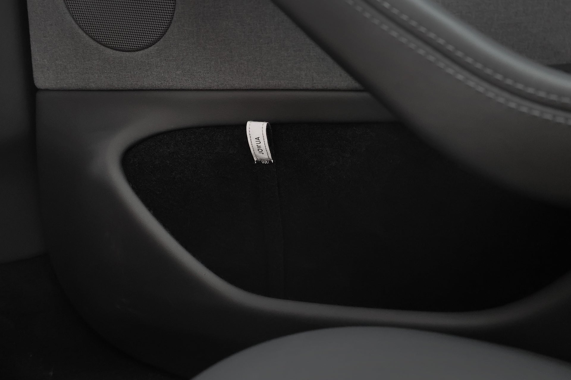 Close-up of a JOWUA Reflective Rear Door Emergency Release Strap (Model 3 Highland, 2024+ / All Model Y) installed on a black car door panel, showing a fabric "AIRBAG" tag and an injection-molded hook near the speaker.